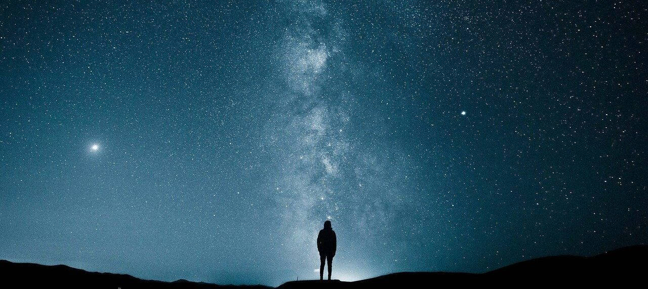 夜空 桌布 壁紙 — 一個孤獨的身影站在山頂上，仰望壯麗的星空，銀河明亮地閃耀。