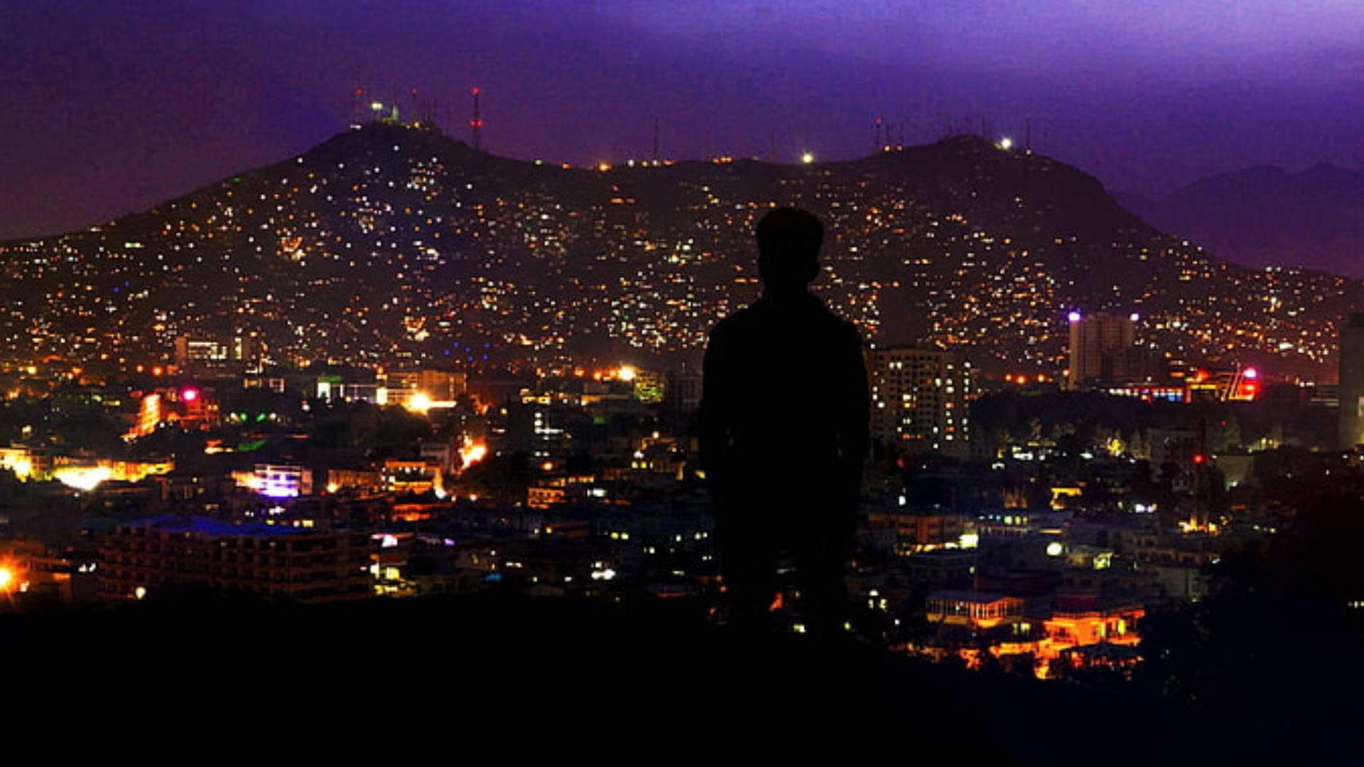 Kabul 桌布 壁紙 — 一個孤獨的身影站在山頂上，俯瞰著在深紫色夜空下，被溫暖燈光照亮的廣闊城市景觀。