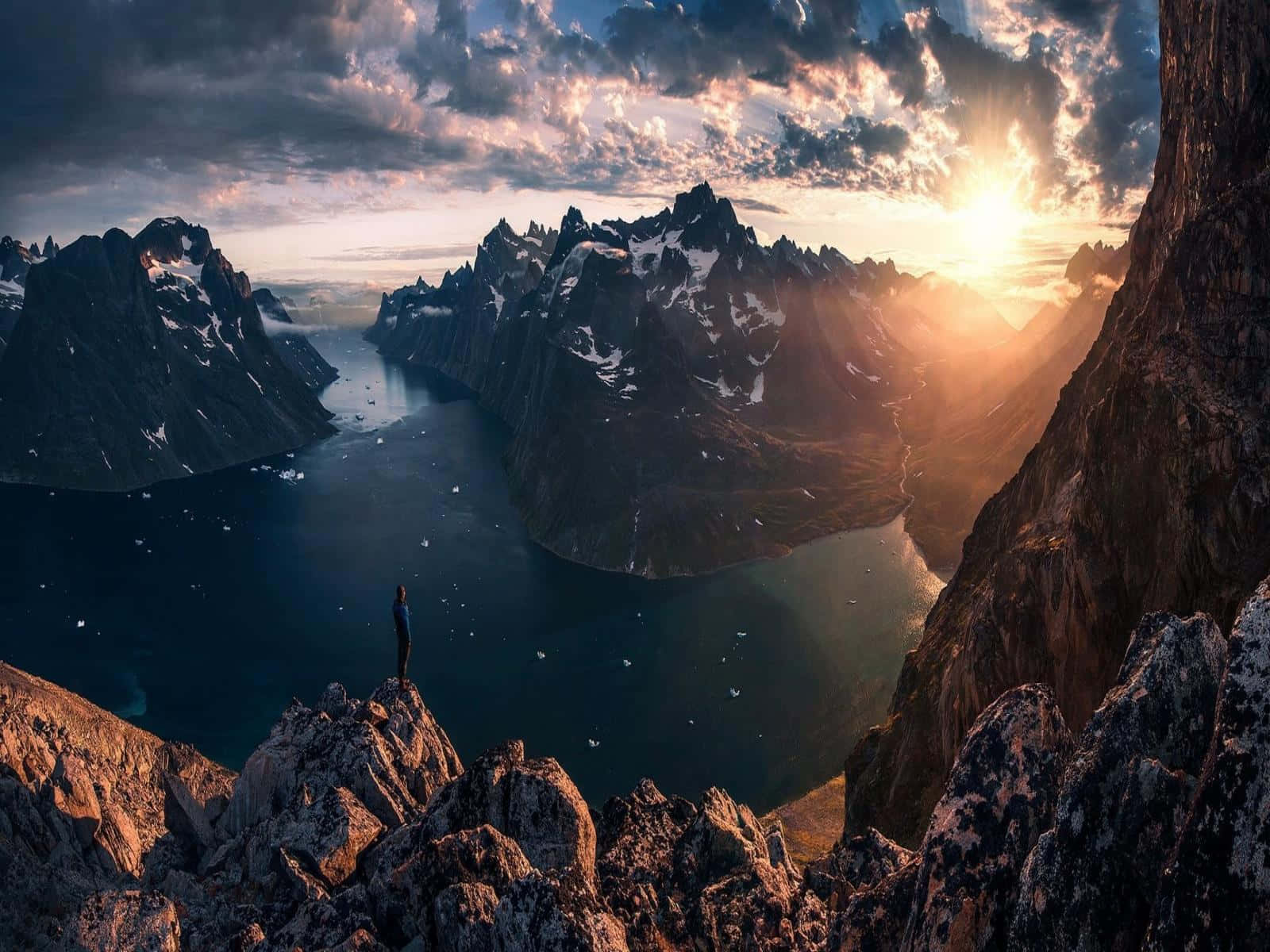 Greenland 桌布 壁紙 — 一個孤獨的身影站在岩石懸崖上，俯瞰著廣闊的峽灣，夕陽在鋸齒狀的雪山後落下。