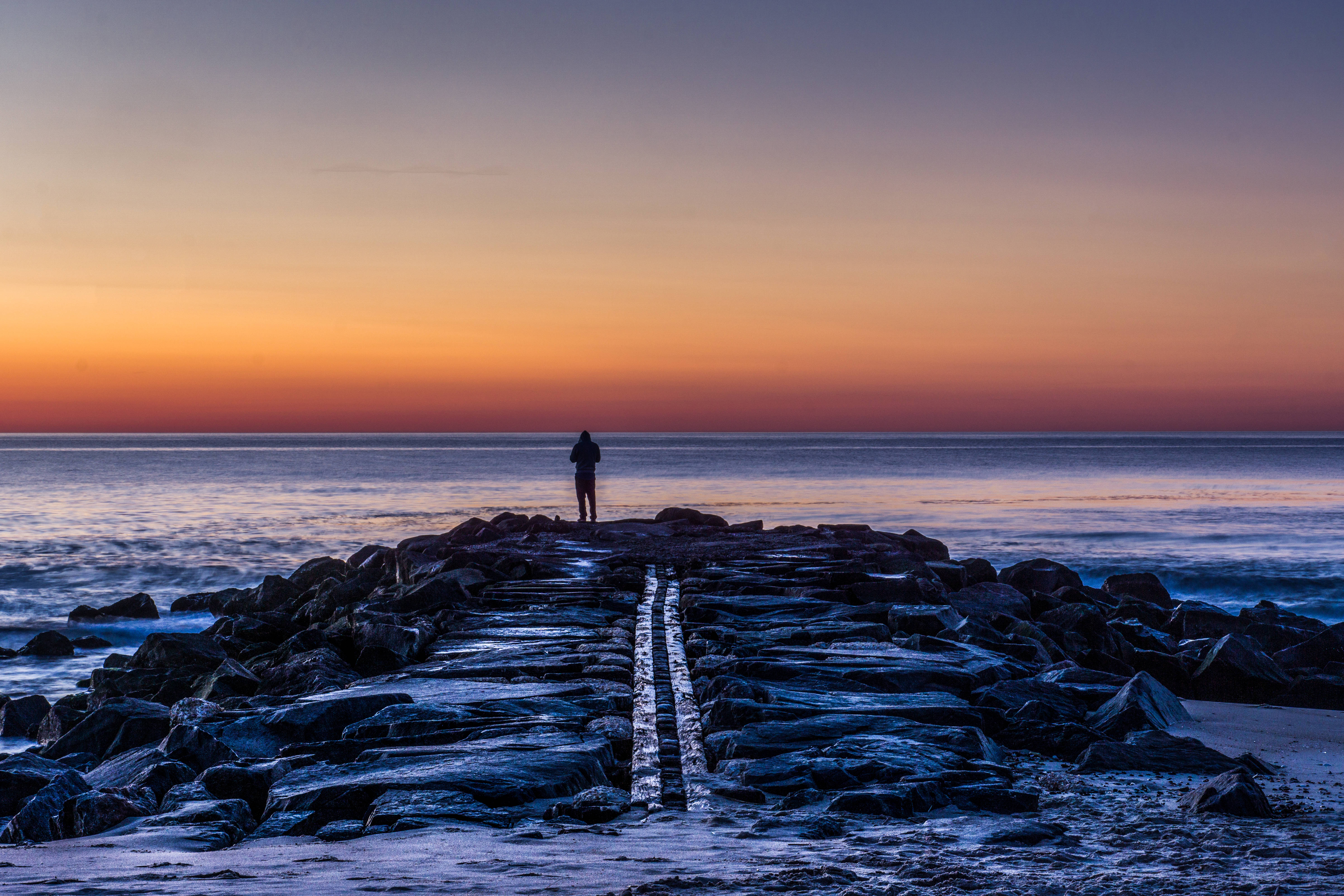 Forever Alone 桌布 壁紙 — 一個孤獨的身影站在岩石碼頭的盡頭，太陽以火熱的橙色和紫色色調在海洋上落下。