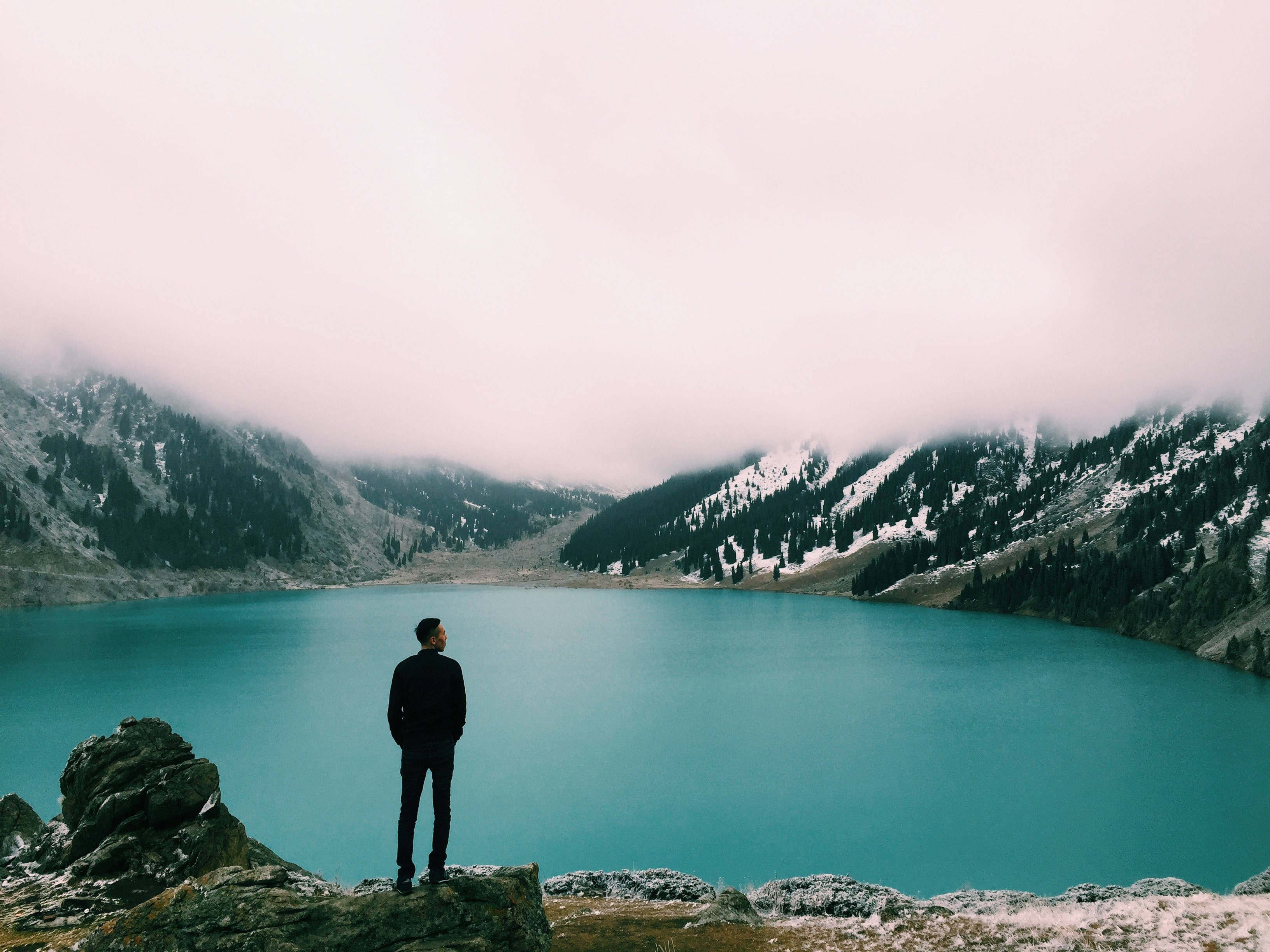 Lake 桌布 壁紙 — 一個孤獨的身影站在岩石露頭上，俯瞰著一個廣闊的綠松石色湖泊，湖泊坐落在被薄霧籠罩的積雪山脈之間。