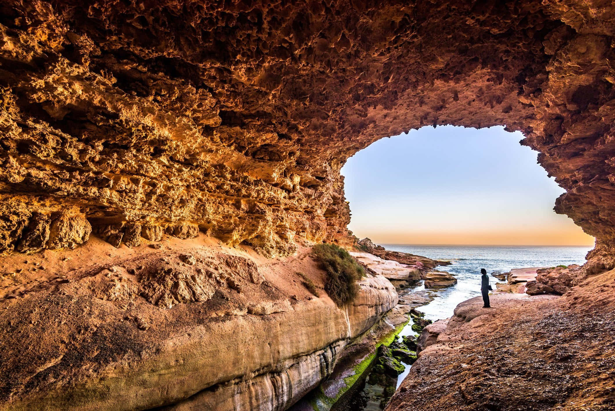 Cave 桌布 壁紙 — 一個孤獨的身影站在崎嶇海岸洞穴的洞口，框住寧靜的海洋日落。