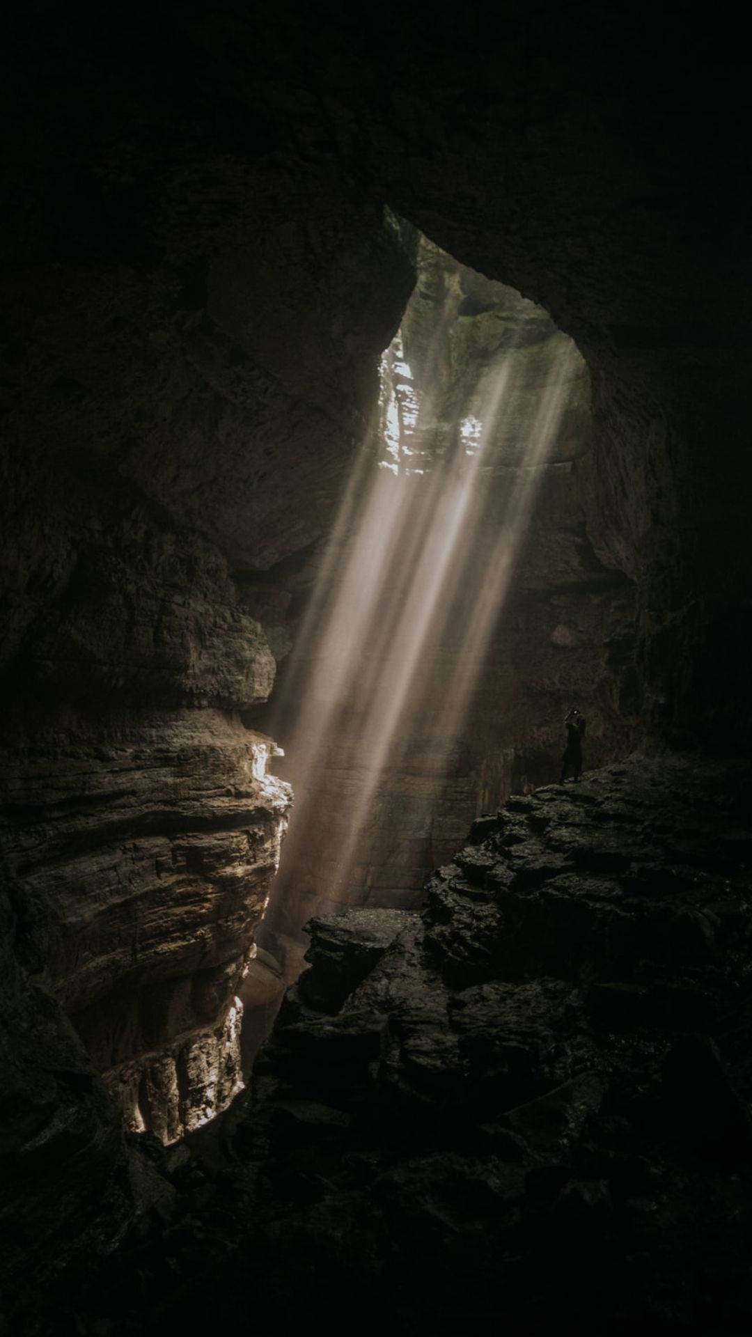 Cave 桌布 壁紙 — 一個孤獨的身影站在廣闊的黑暗洞穴中，神聖的陽光從上方的開口穿透進來，照亮了崎嶇的岩石 formations。