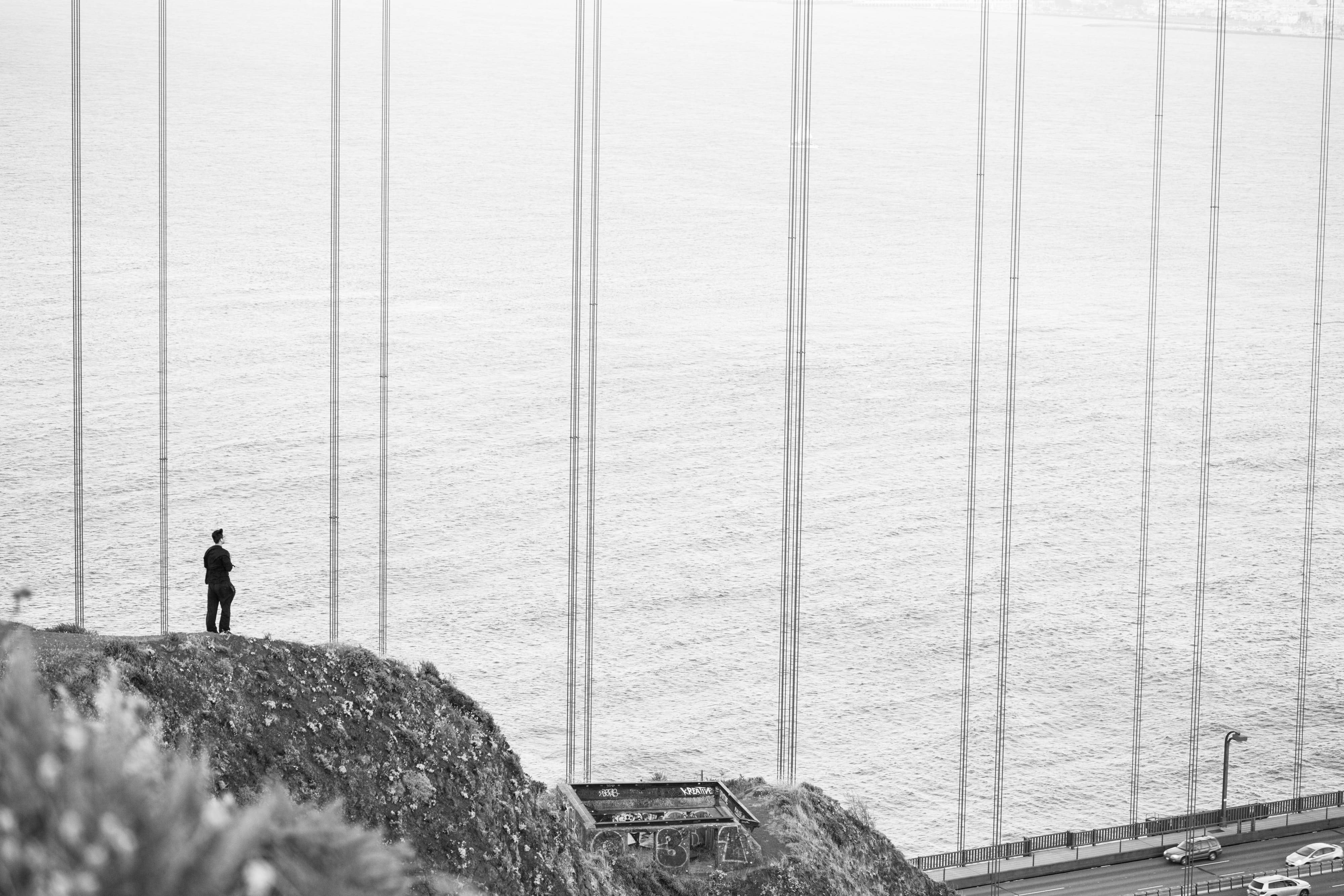 Alone 桌布 壁紙 — 一個孤獨的身影站在懸崖上，俯瞰著廣闊的水面和橋樑高聳的懸索，以單色呈現。