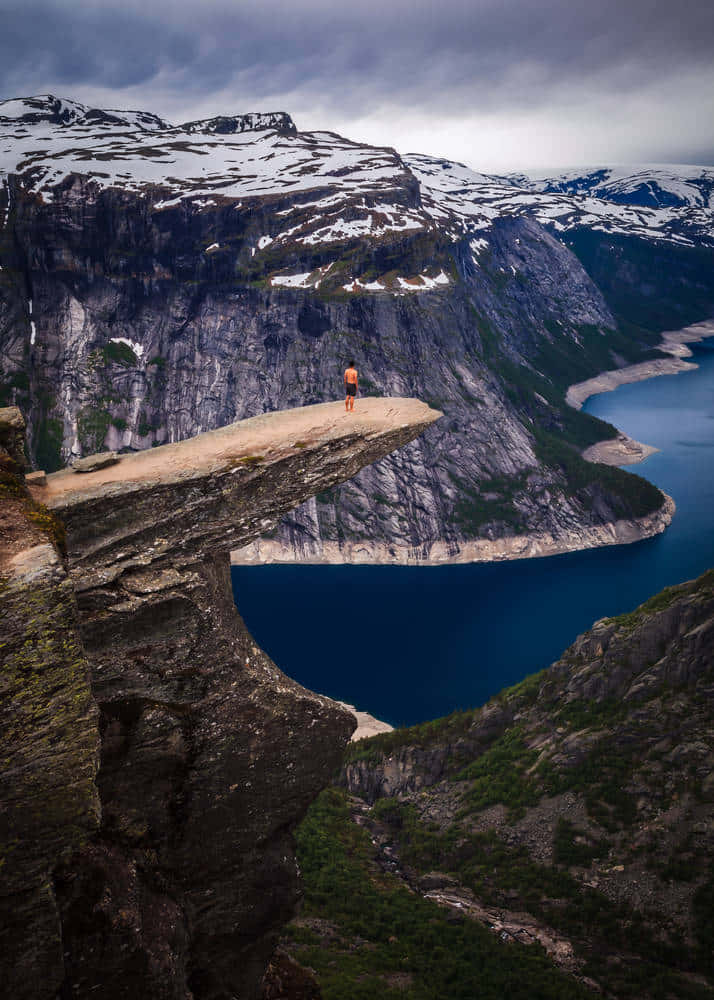 Norway 桌布 壁紙 — 一個孤獨的身影站在戲劇性的Preikestolen懸崖頂上，俯瞰著深藍色的峽灣和白雪覆蓋的山脈，天空多雲。
