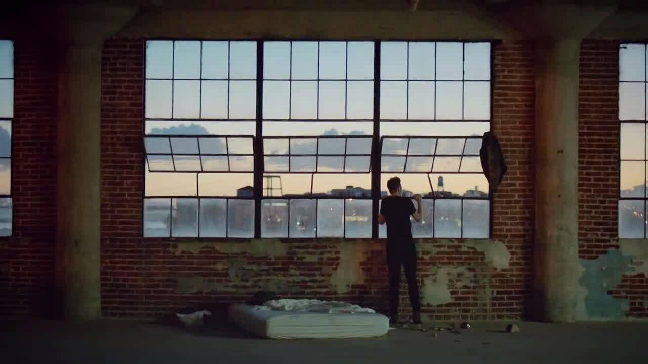 Bastille 桌布 壁紙 — 一個孤獨的身影站在昏暗的磚造倉庫中，透過一扇大破窗凝視著黃昏時分的城市天際線。