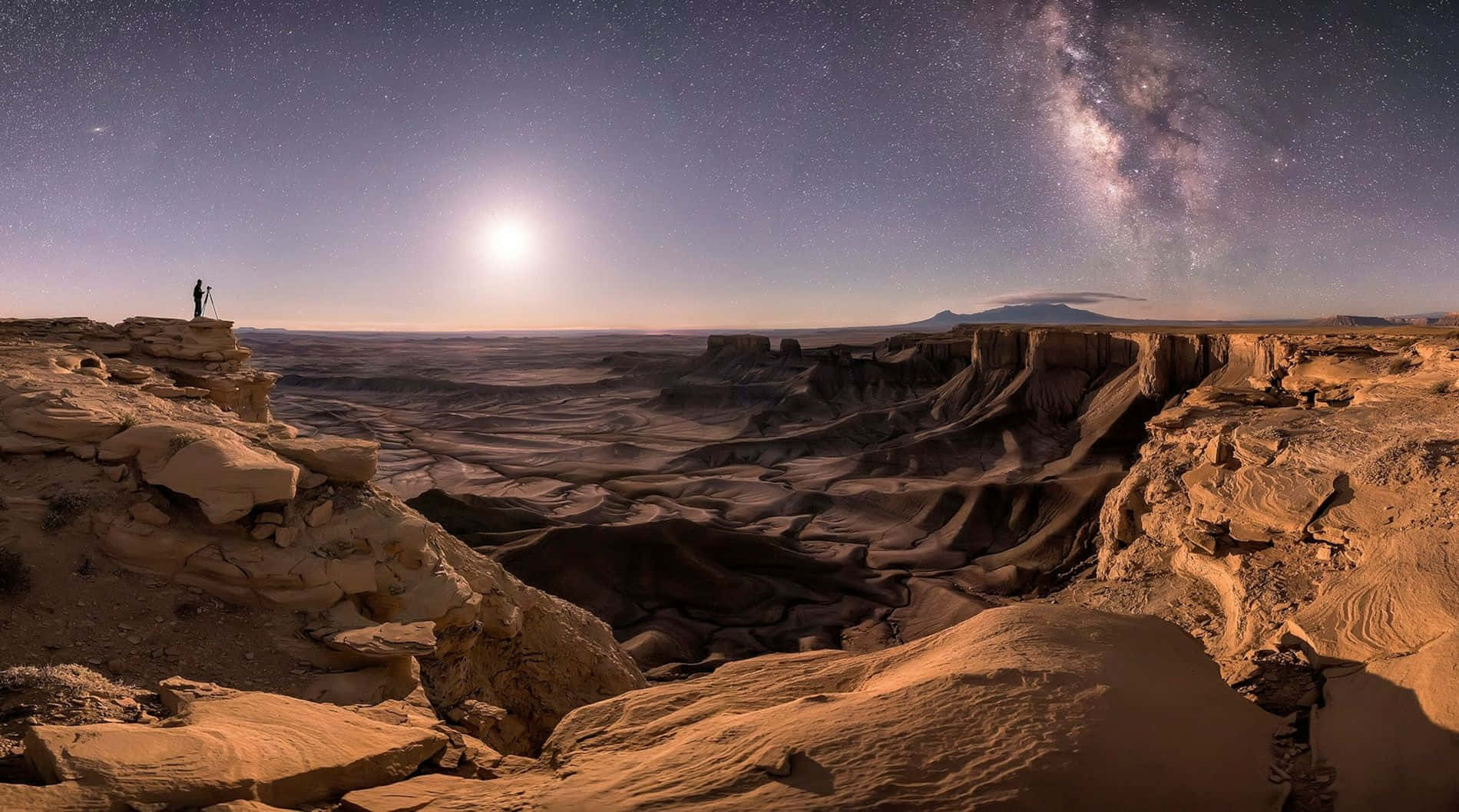 astronomy 桌布 壁紙 — 一個孤獨的身影站在沙漠懸崖上，夜晚星空下，銀河與明亮的月亮照亮下方崎嶇的峽谷。