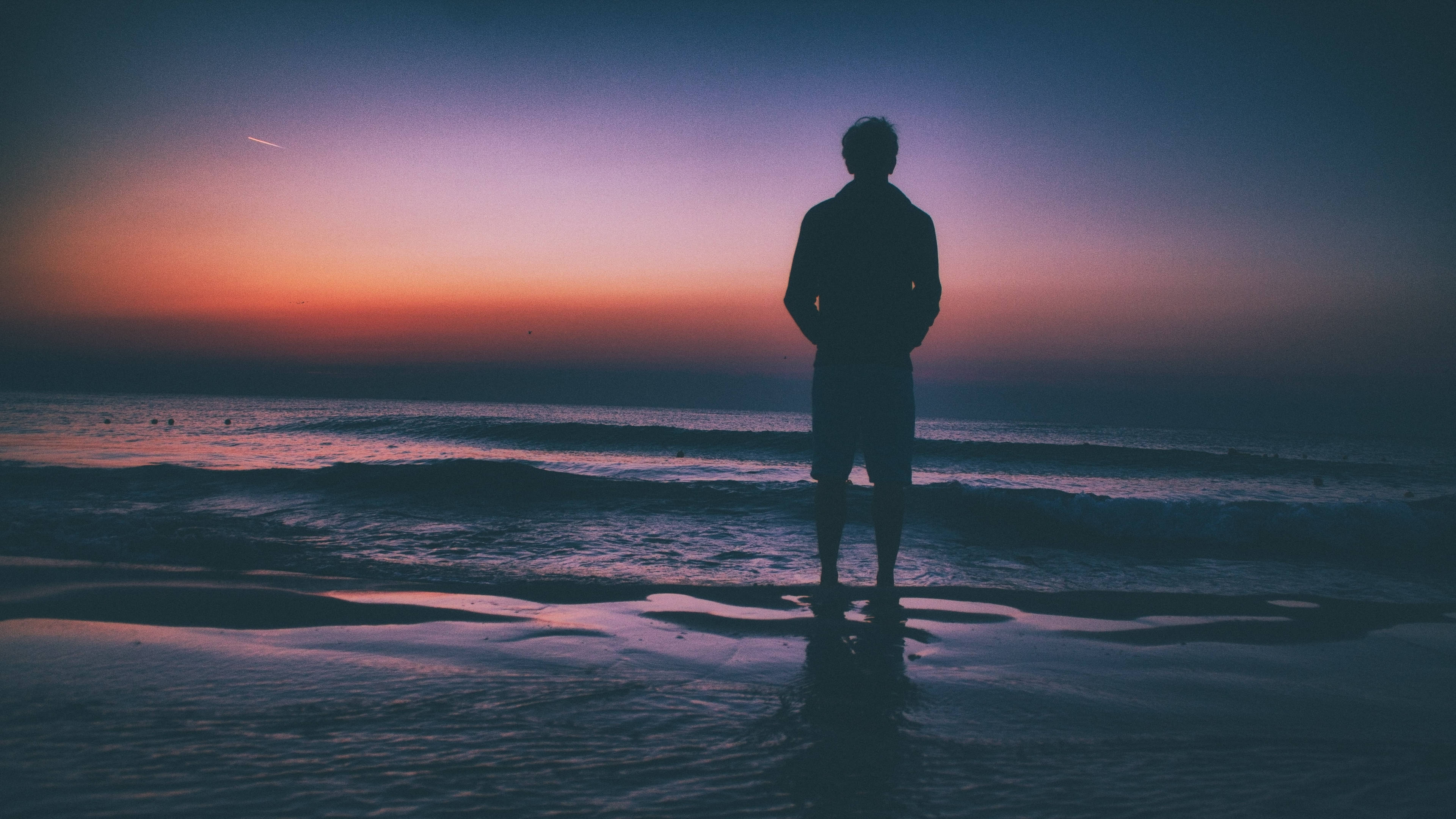 Alone Boy 桌布 壁紙 — 一個孤獨的身影站在海岸線上，海浪在紫、橙、深藍色調的暮色天空下拍打。