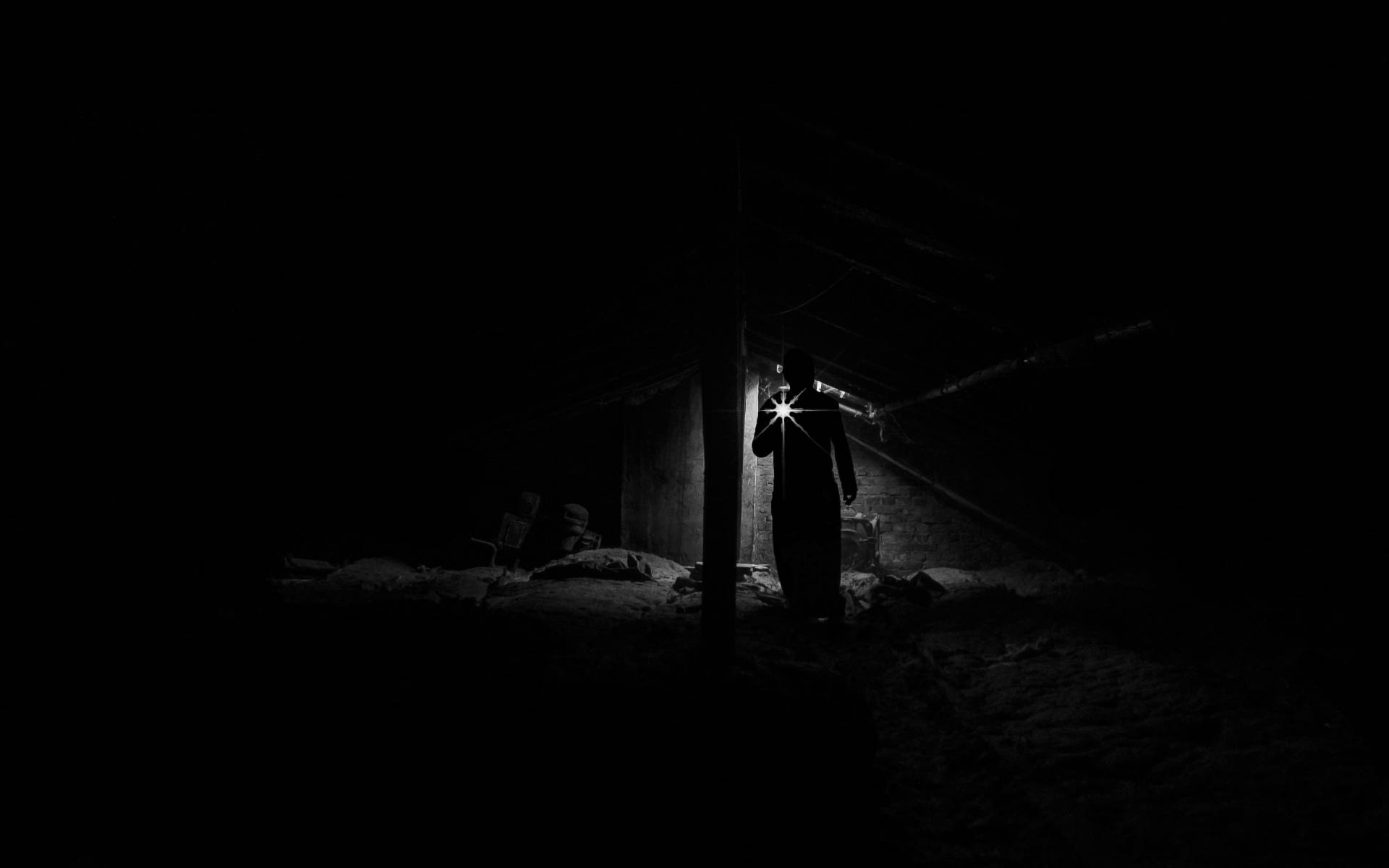 Alone 桌布 壁紙 — 一個孤獨的身影站在漆黑的閣樓中，手持明亮的手電筒，劃破黑暗，照亮空氣中的灰塵微粒。