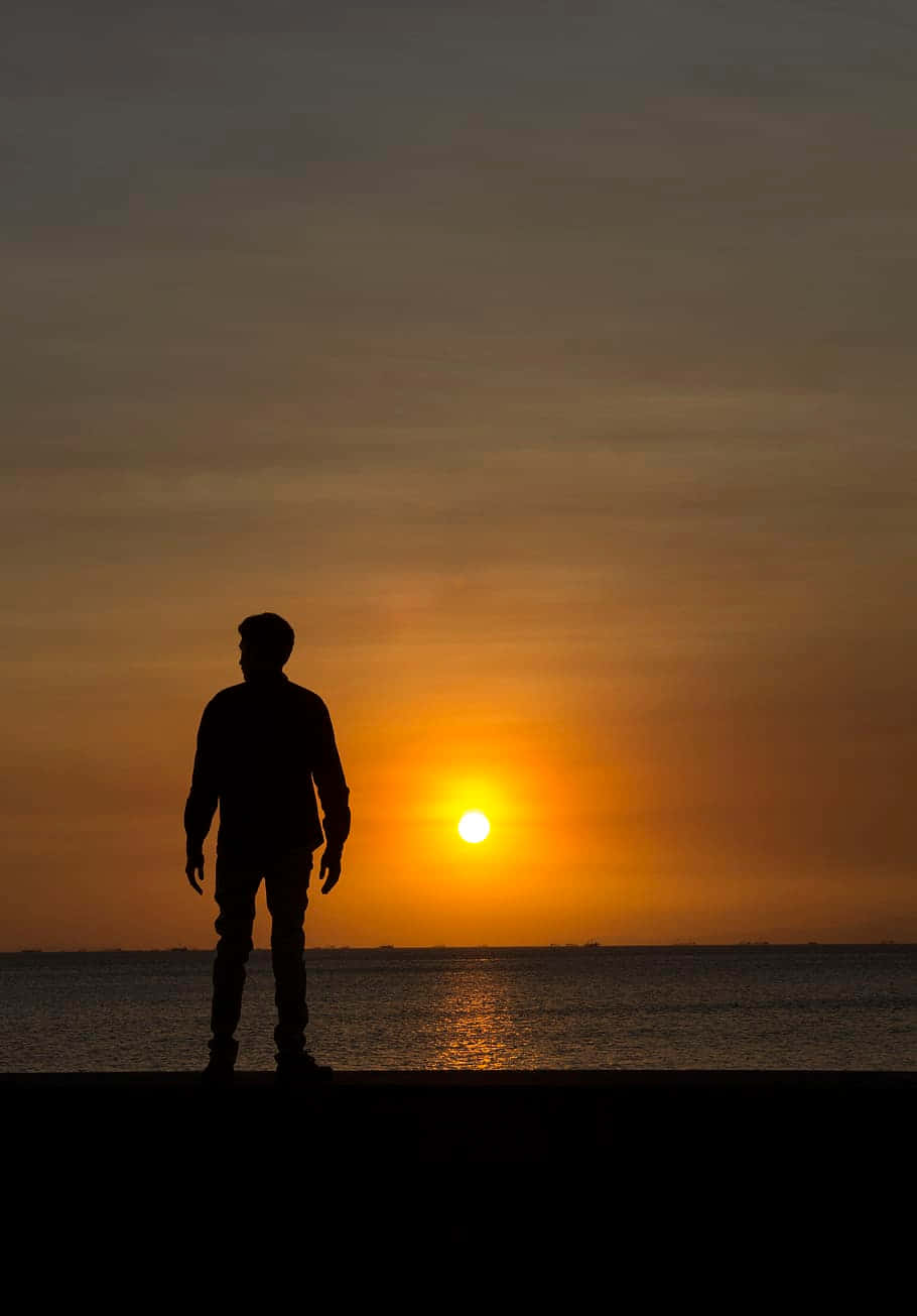Silhouette 桌布 壁紙 — 一個孤獨的身影站在碼頭上，看著夕陽在平靜的水面上落下，將金色的光芒灑向天空。