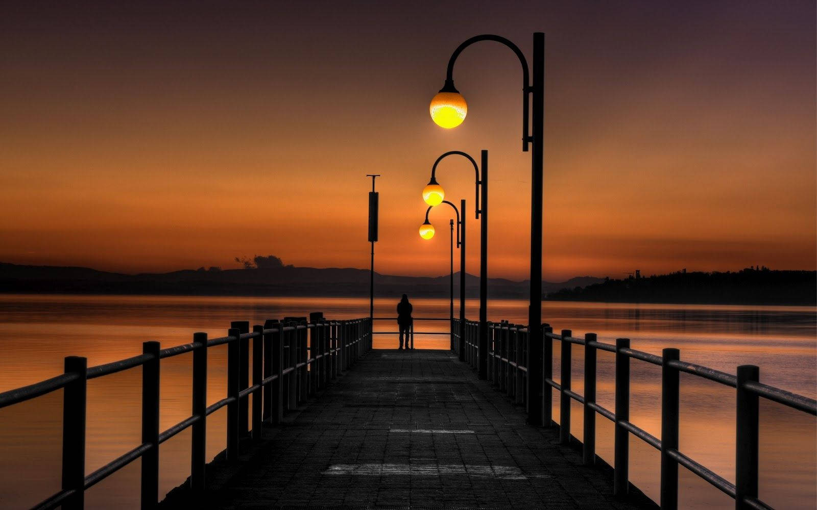 Forever Alone 桌布 壁紙 — 一個孤獨的身影站在碼頭盡頭，頭頂是發光的街燈，天空在日落時燃燒成橙色和紫色。