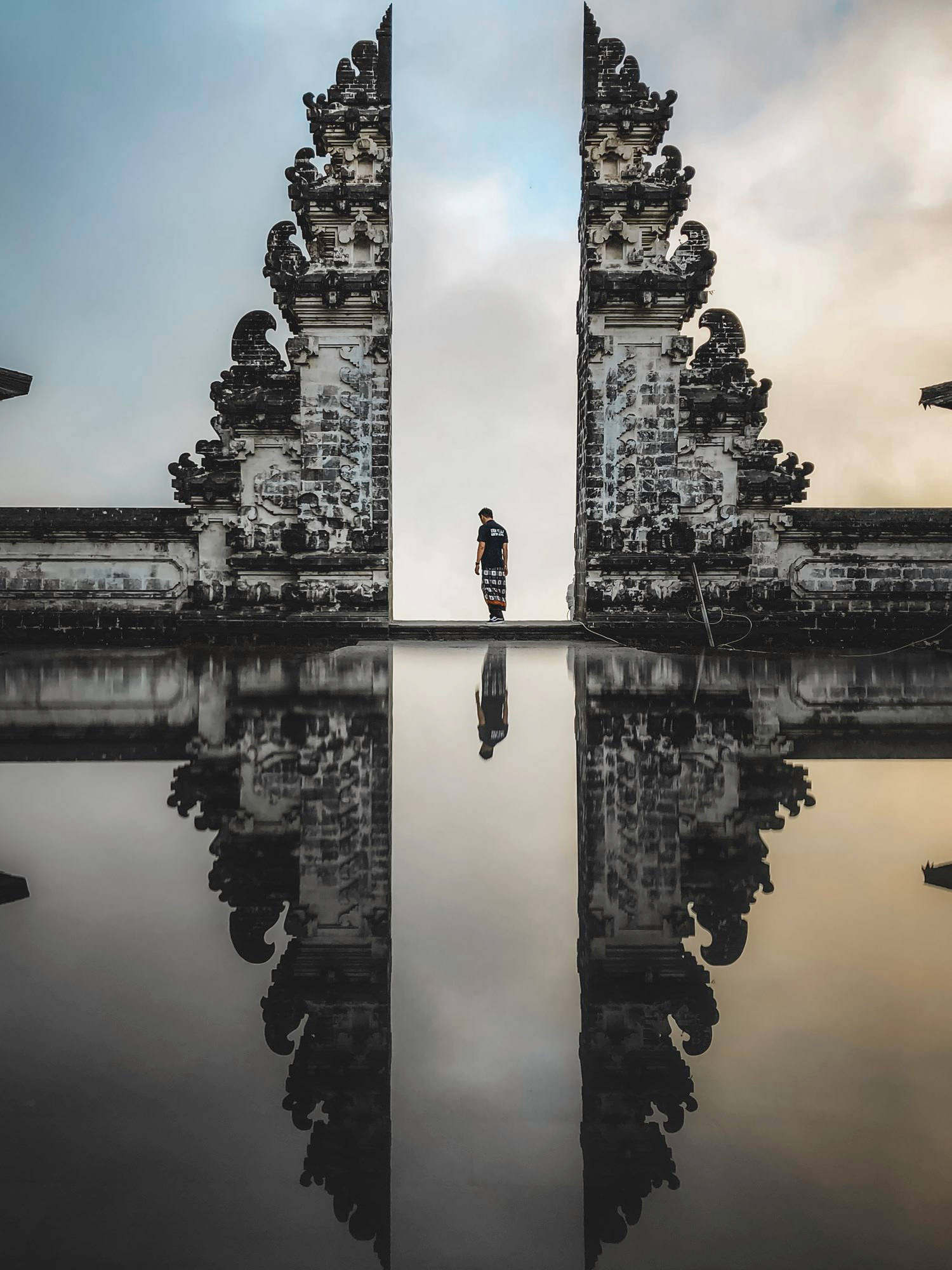 Travel And Tourism 桌布 壁紙 — 一個孤獨的身影站在裝飾性石門之間，在平靜的水面上完美倒映，天空柔和。