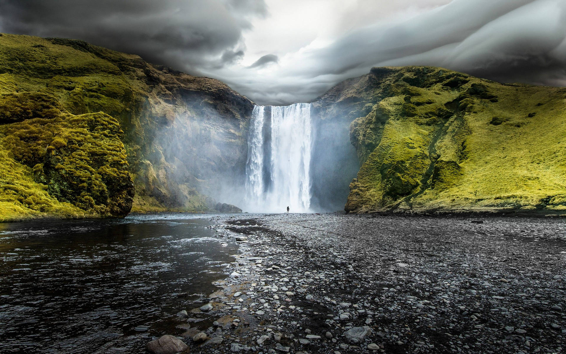 Iceland 桌布 壁紙 — 一個孤獨的身影站在雄偉的瀑布前，瀑布在長滿苔蘚的綠色懸崖之間傾瀉而下，天空烏雲密布，氣氛戲劇化。