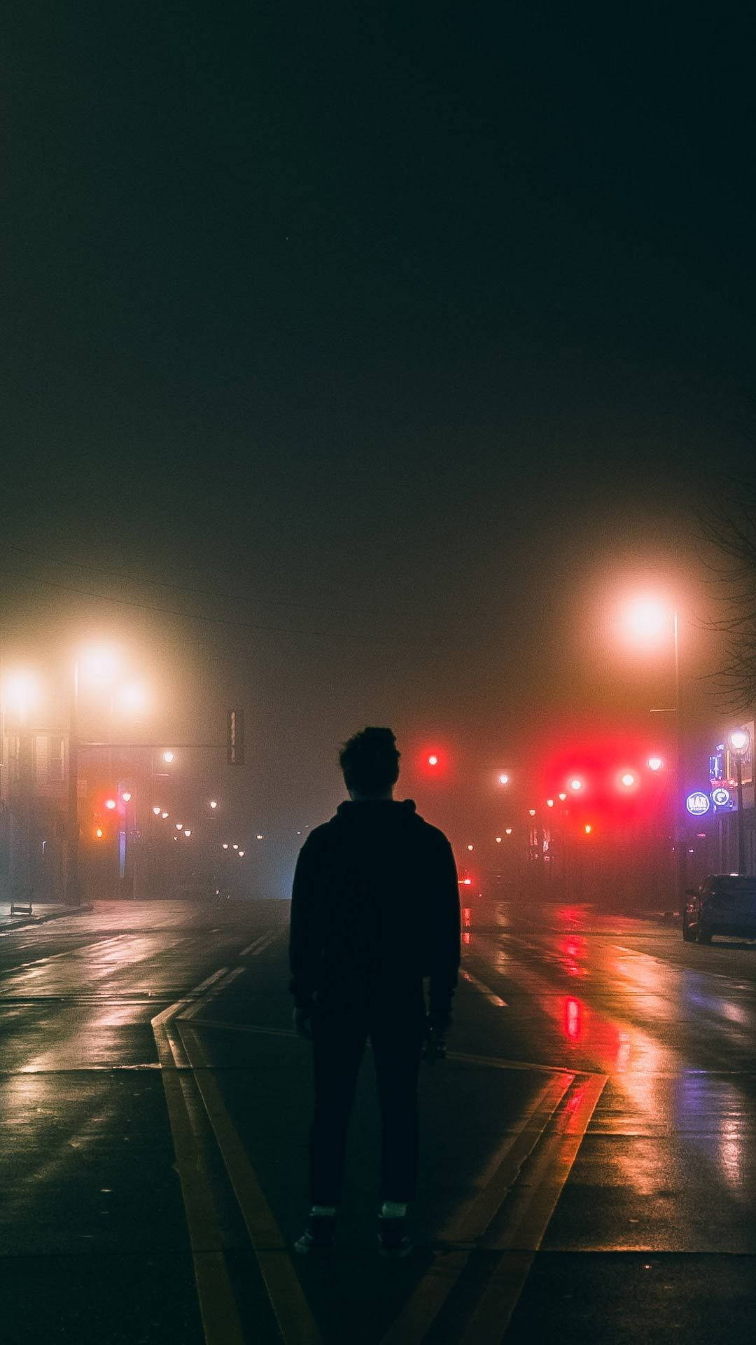 Best Dark 桌布 壁紙 — 一個孤獨的身影站在霧氣瀰漫的潮濕街道中央，夜晚被發光的街燈和紅色交通信號燈照亮。
