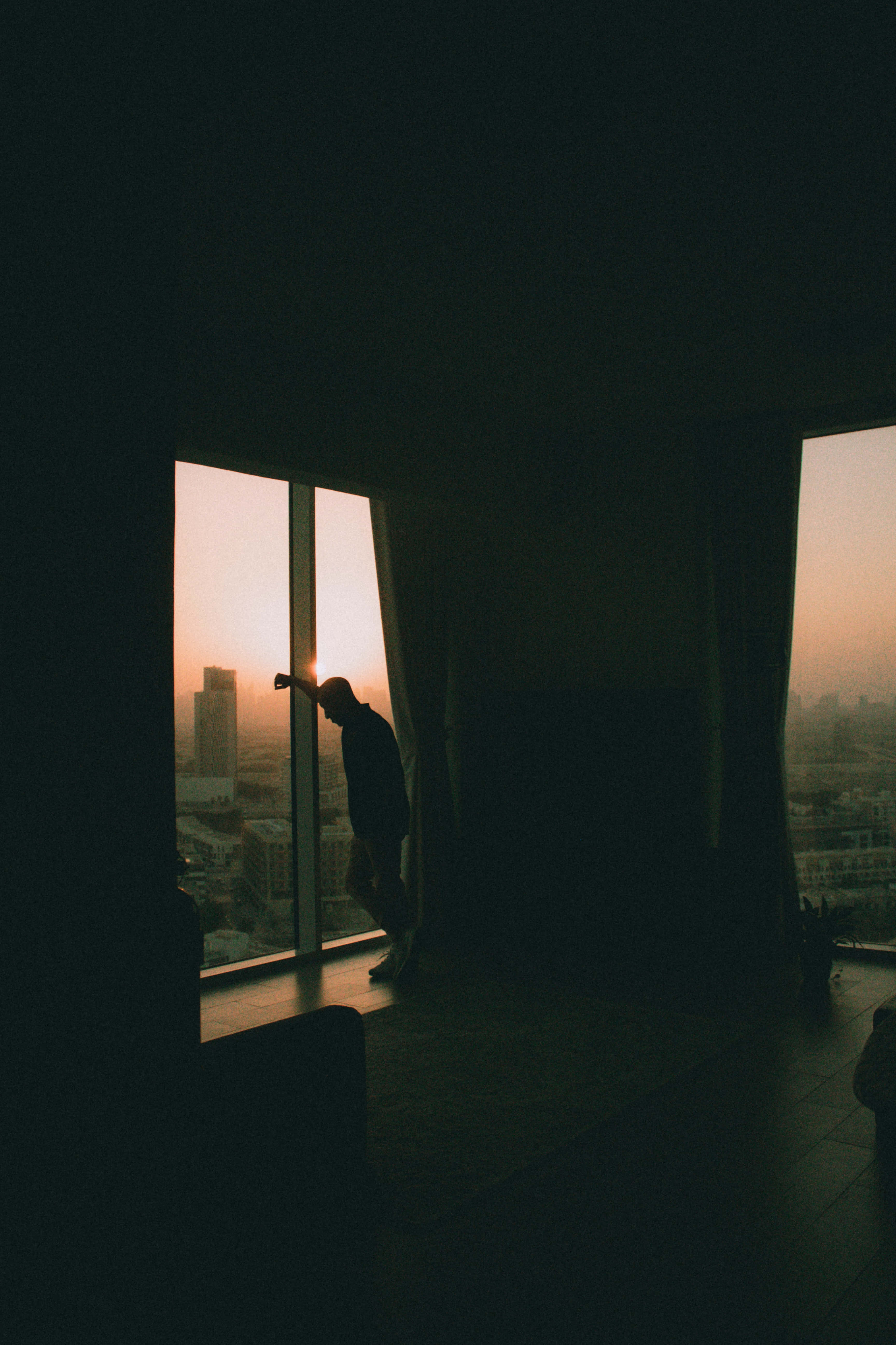 Aesthetic Room 桌布 壁紙 — 一個孤獨的身影站在高層窗邊，捕捉沐浴在溫暖夕陽餘暉中的城市景觀。