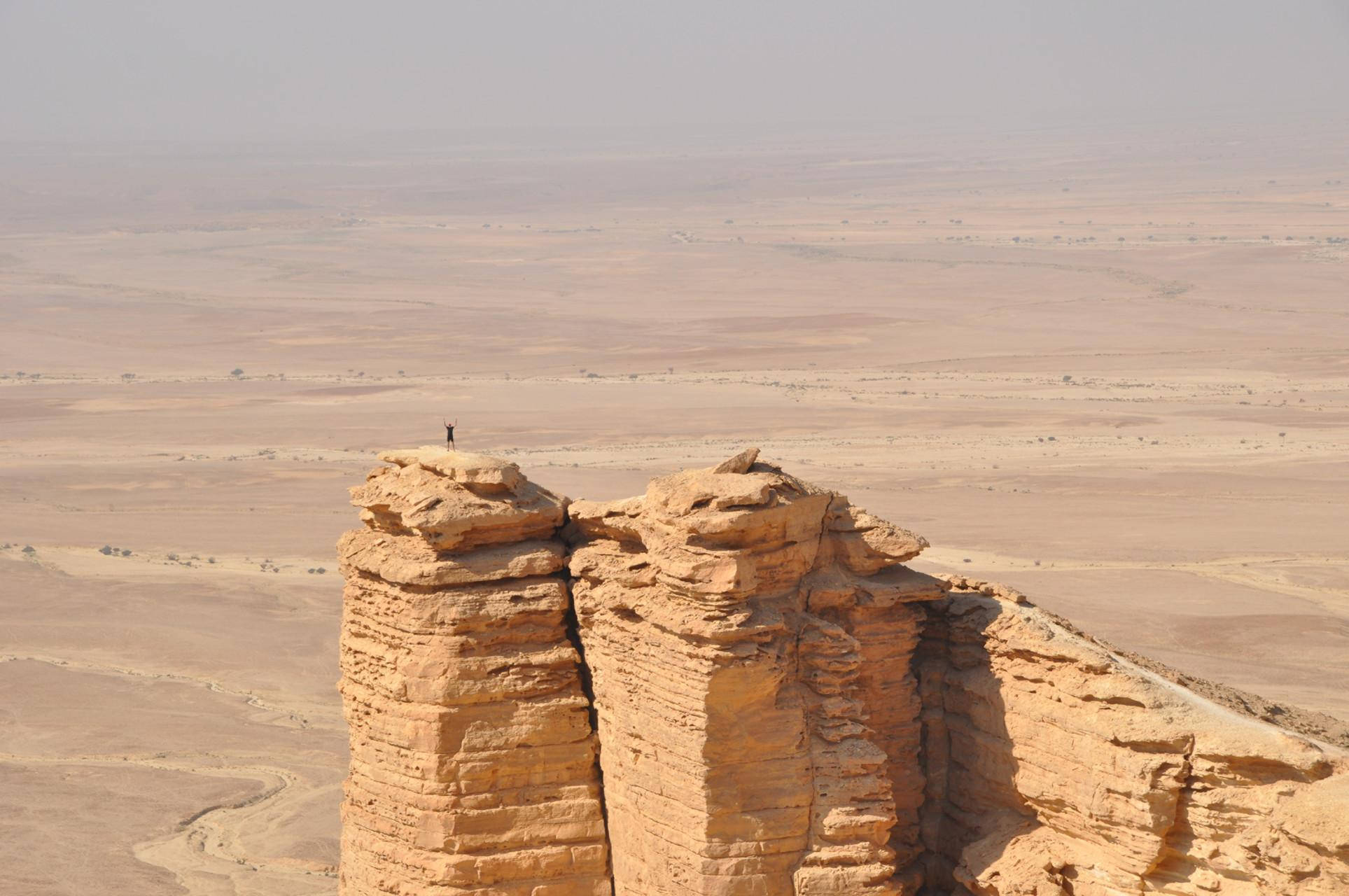 Saudi 桌布 壁紙 — 一個孤獨的身影站在高聳的橙色砂岩懸崖頂上，俯瞰著朦朧日光下無邊無際的乾旱沙漠平原。