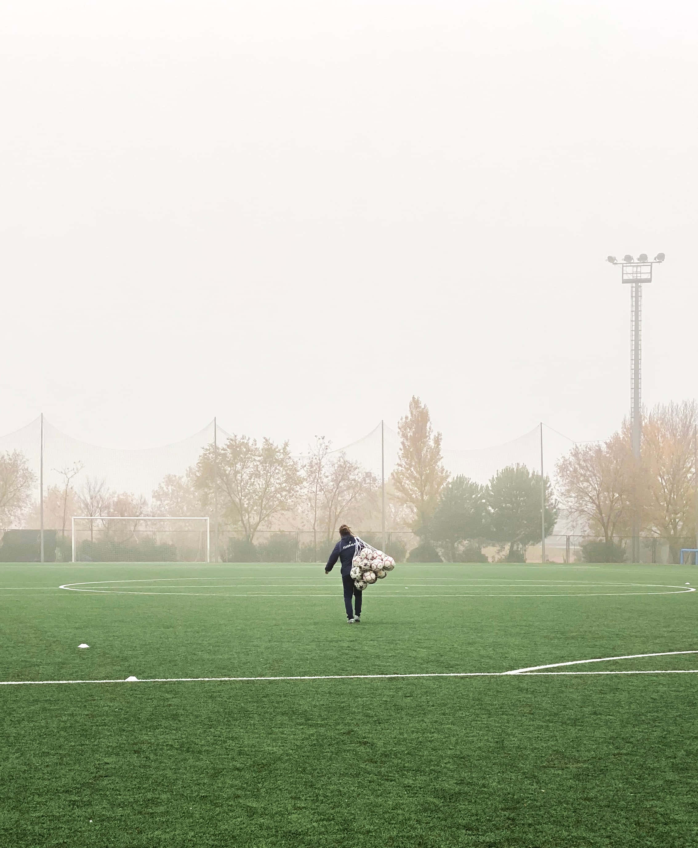 Soccer Ball 桌布 壁紙 — 一個孤獨的身影背著一捆足球，穿過霧氣瀰漫的綠色球場，樹木和球門在霧中逐漸消失。