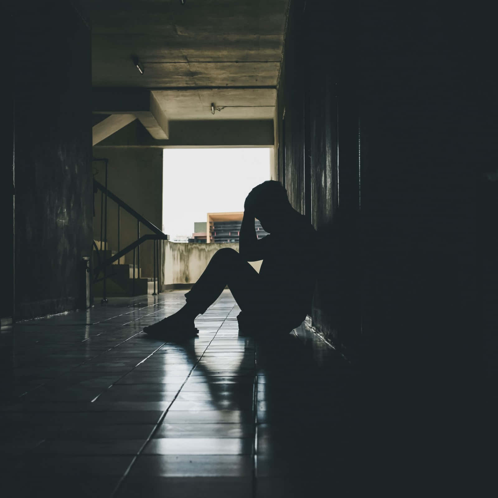 Depressed 桌布 壁紙 — 一個孤獨的身影蜷縮在昏暗走廊的瓷磚地上，雙手抱頭，被敞開門口射入的強烈背光所籠罩。