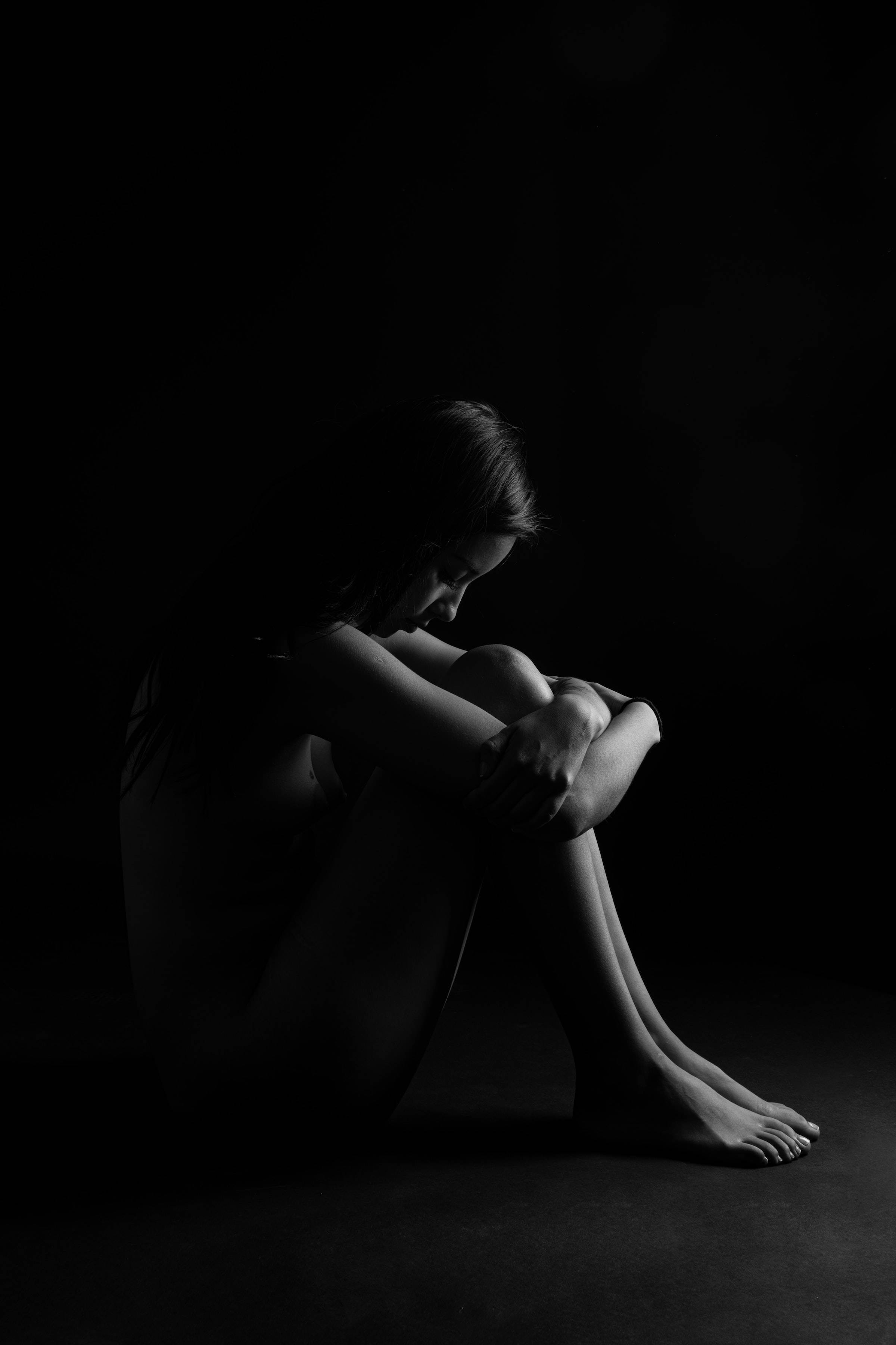 Woman 桌布 壁紙 — 一個孤獨的身影蜷縮在陰影中，低垂著頭，被一道戲劇性的光線照亮，背景是一片漆黑的虛空。