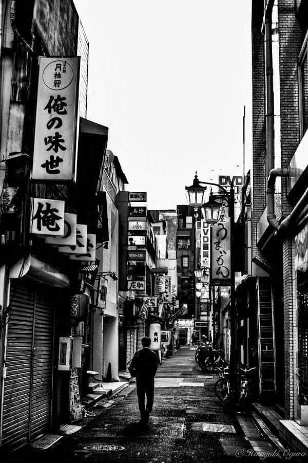 黑色美學 桌布 壁紙 — 一個孤獨的身影走在一條狹窄、陰暗的日本小巷中，兩旁是傳統招牌和關閉的商店，天空晴朗。