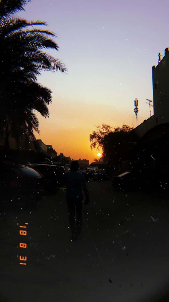 Chill Aesthetic 桌布 壁紙 — 一個孤獨的身影走在棕櫚樹成蔭的街道上，夕陽落在建築物後面，投下長長的影子。