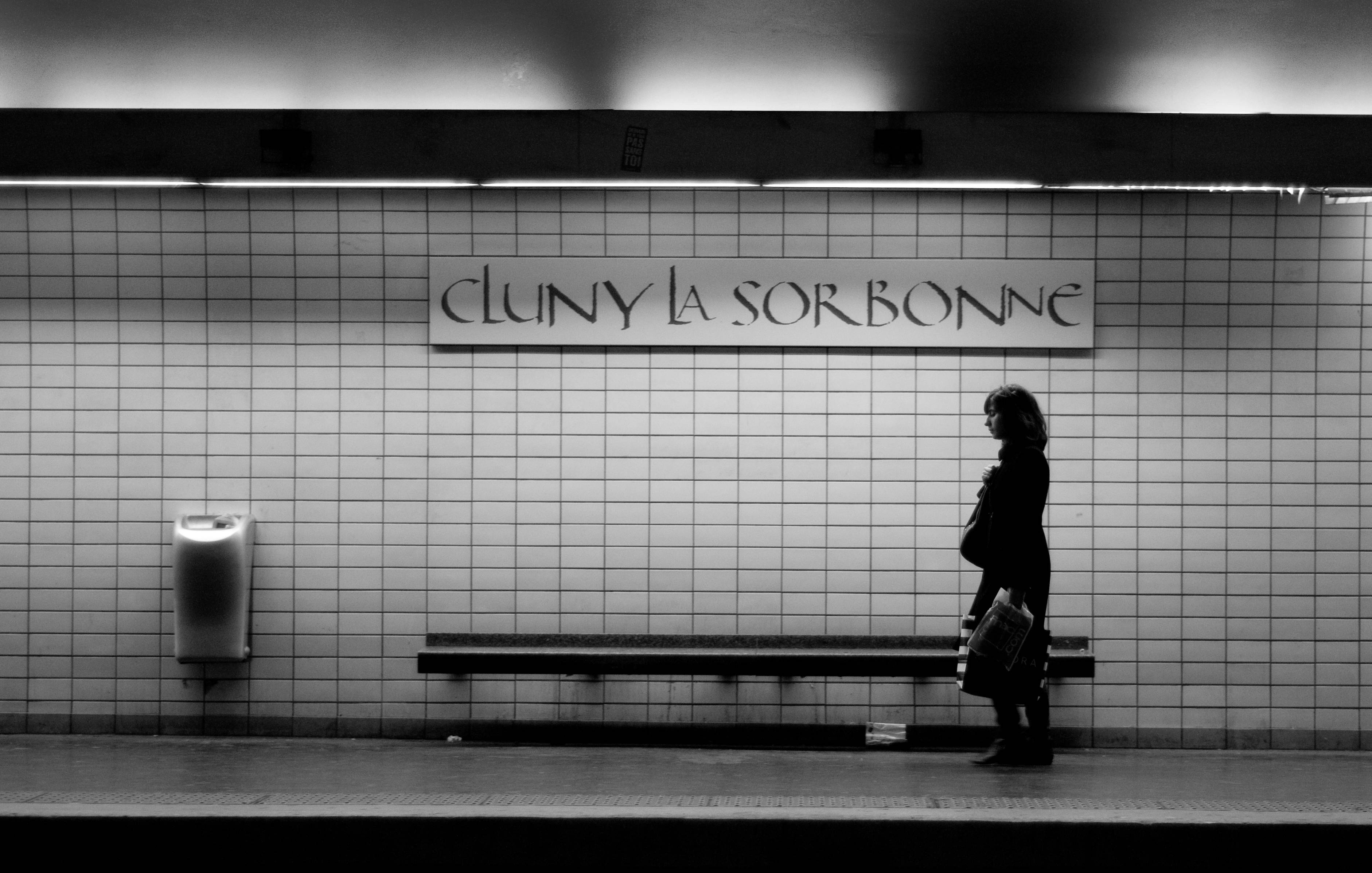 Subway 桌布 壁紙 — 一個孤獨的身影走過一張長椅，下方是「Cluny La Sorbonne」標誌，身處簡樸的磁磚地鐵站。
