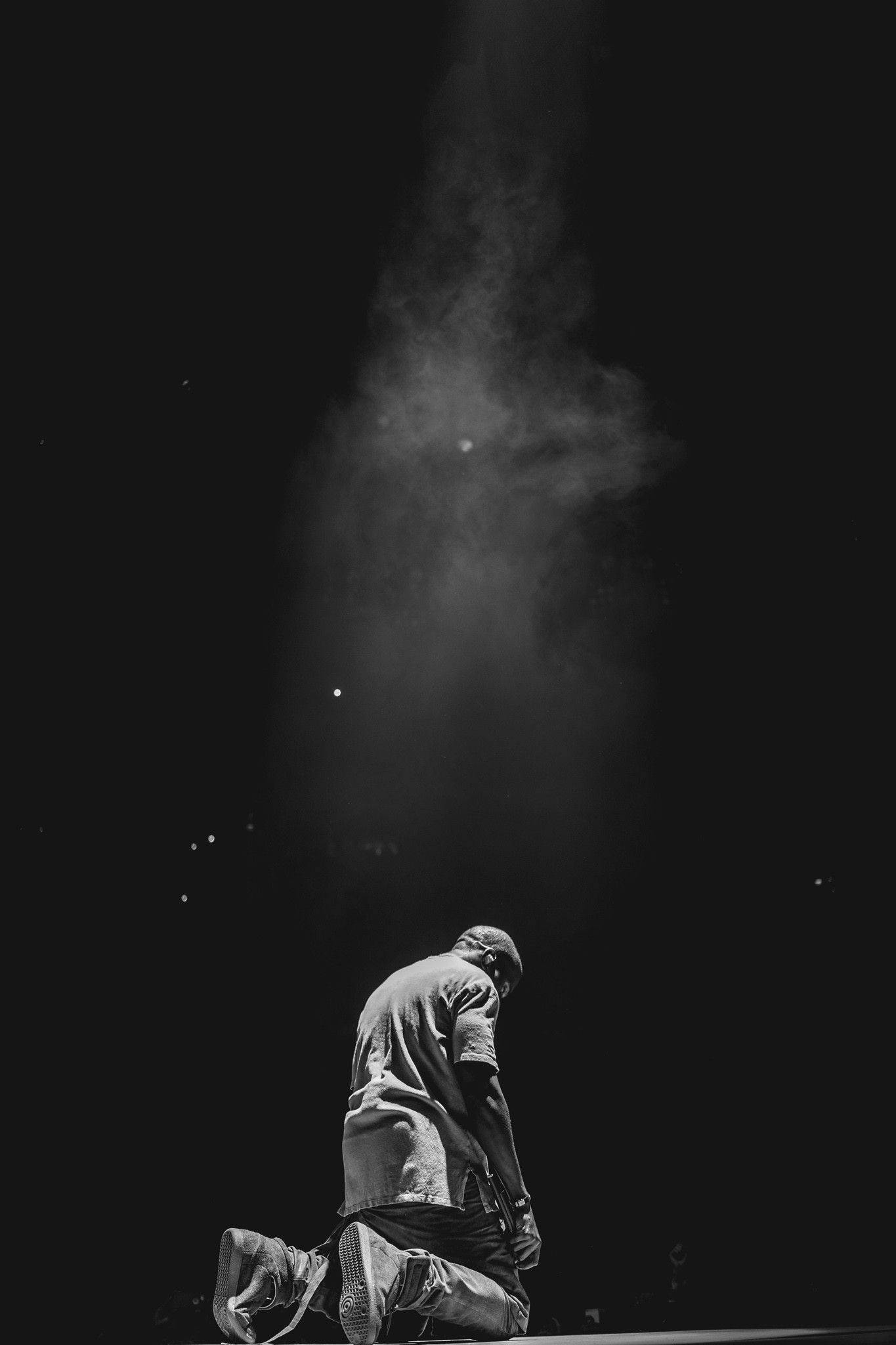 Kanye 桌布 壁紙 — 一個孤獨的身影跪在黑暗中，一道戲劇性的光束照亮了上方的灰塵顆粒。