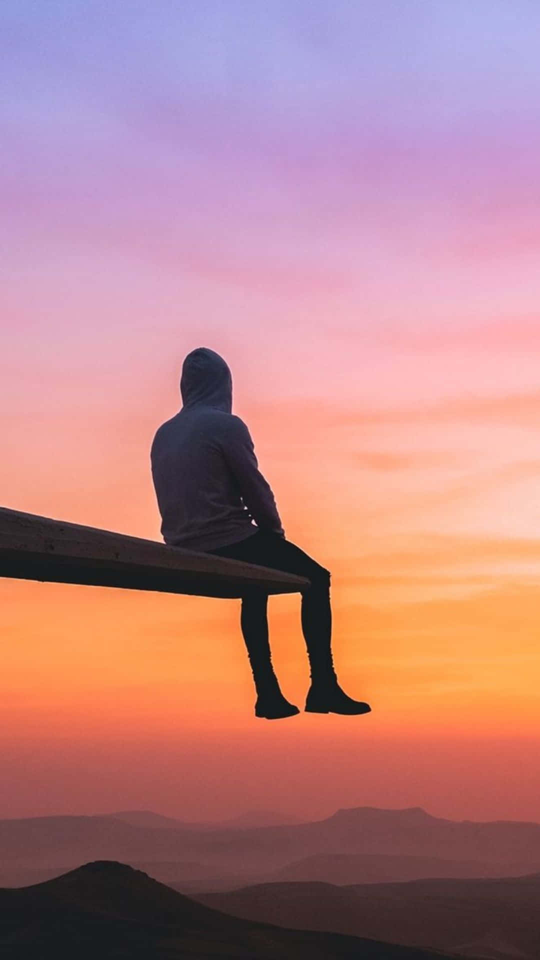Calm Aesthetic 桌布 壁紙 — 一個孤獨的連帽人物坐在木長椅上，俯瞰日落時分的山脈，天空呈現鮮豔的橙色和紫色。