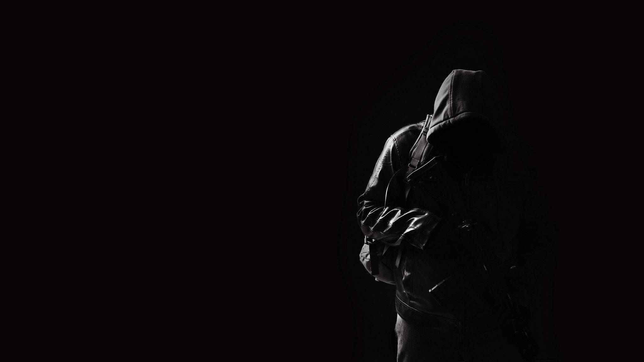 Hoodie 桌布 壁紙 — 一個孤獨的連帽人物穿著皮夾克，站在近乎黑暗的環境中，只有微弱的光線勾勒出他們的輪廓。