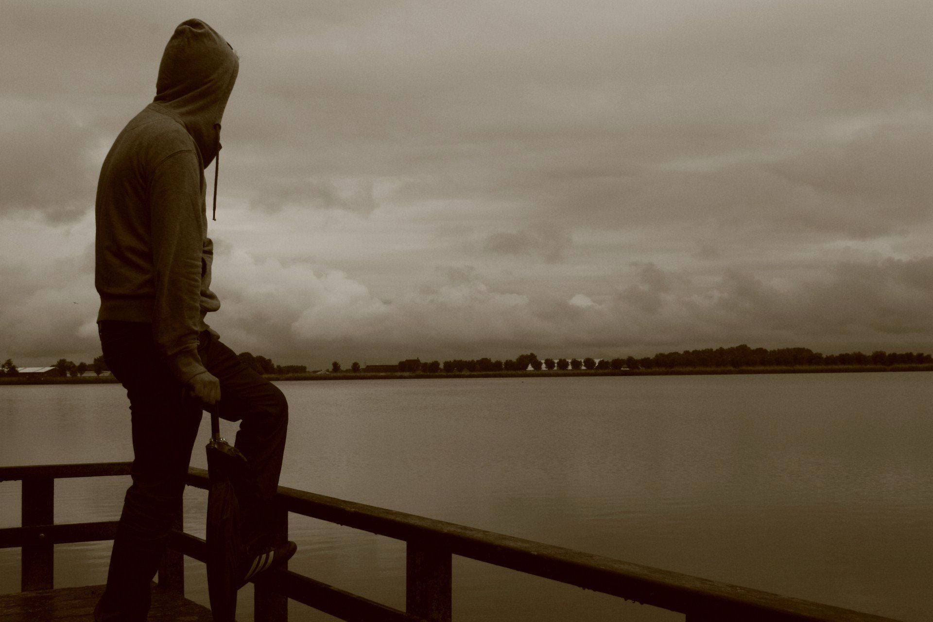 Hoodie 桌布 壁紙 — 一個孤獨的連帽身影坐在木棧橋上，沉思地望著陰天下一片廣闊平靜的湖泊。