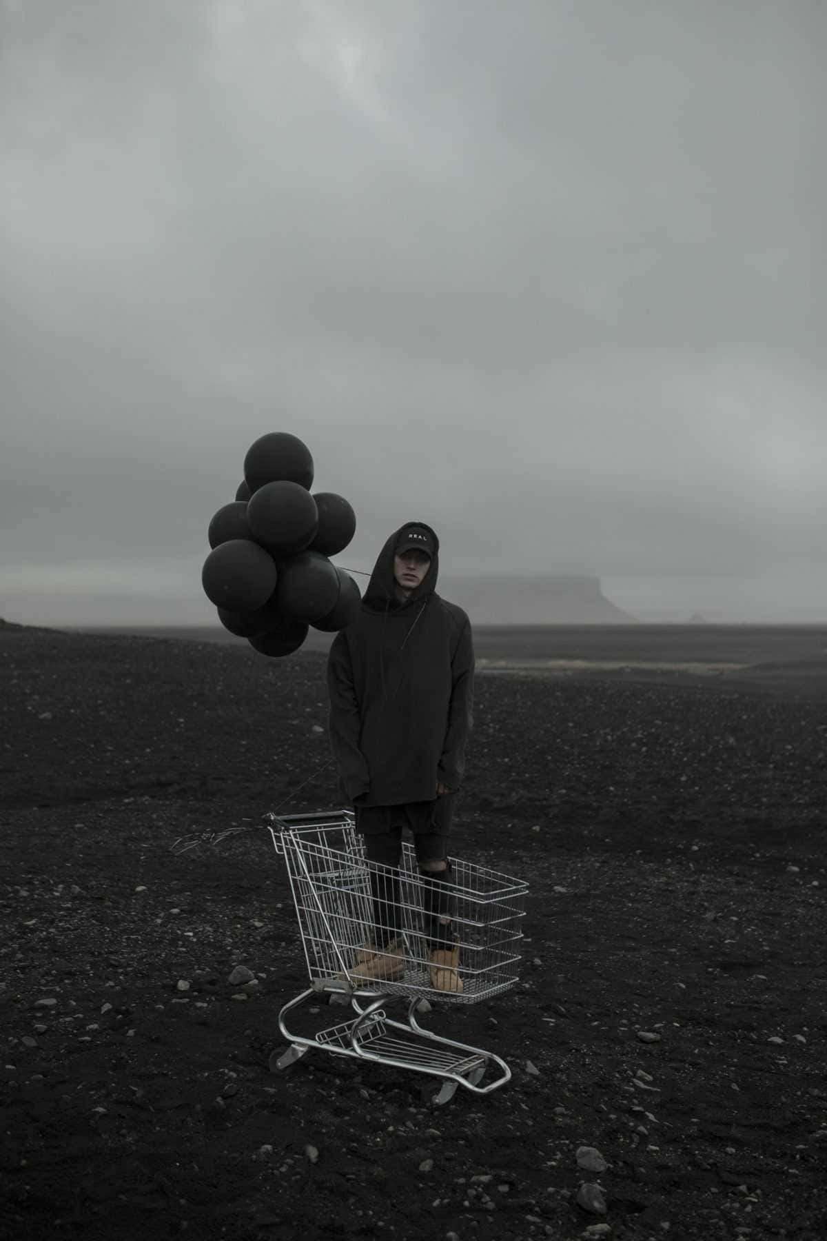 Nf Rapper 桌布 壁紙 — 一個孤獨的連帽身影站在荒涼的黑色熔岩地上，在灰色天空下，於一架被遺棄的購物車旁手持黑色氣球。