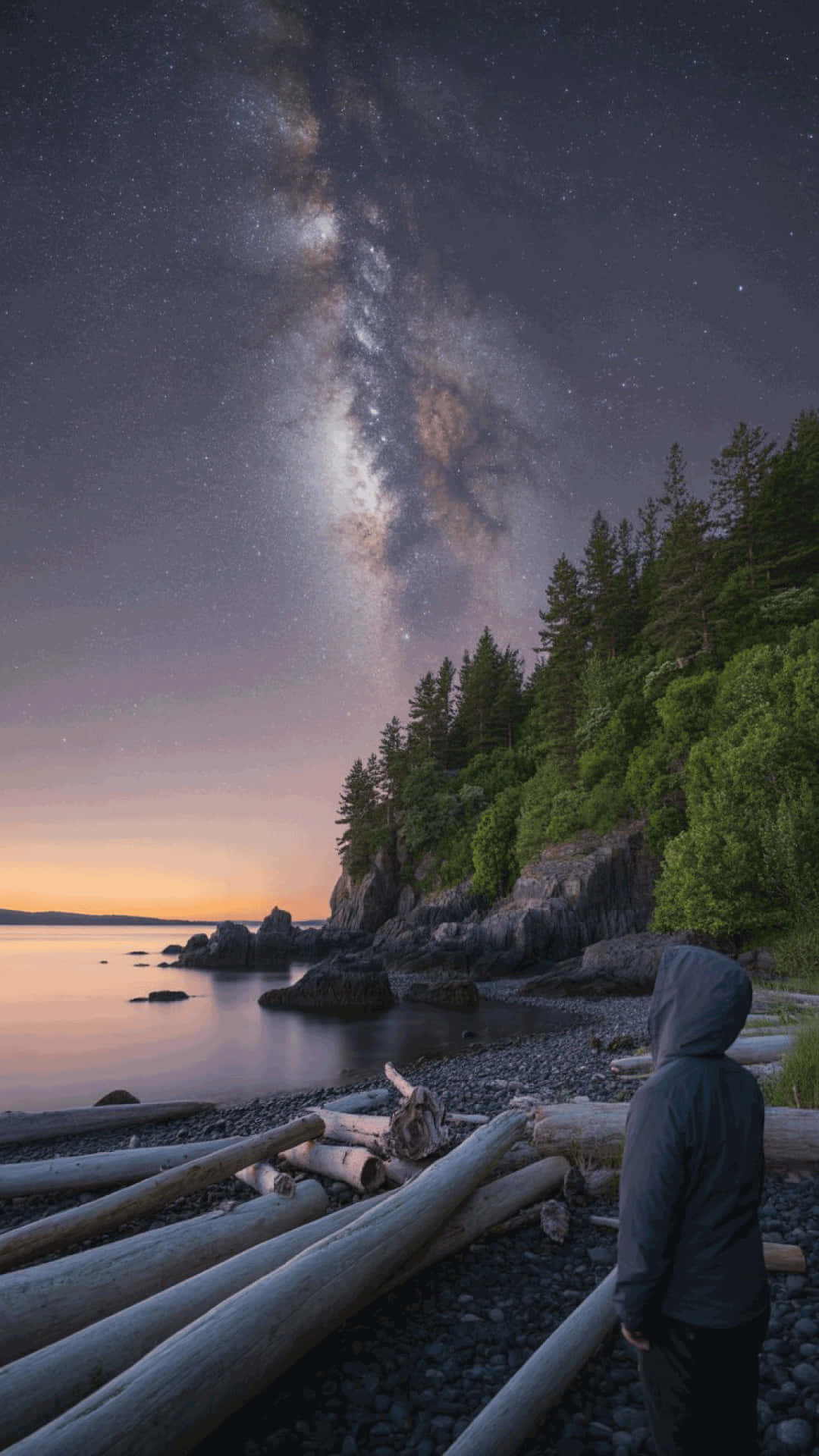 Night sky and seascape at cape serenity milky way wallpaper 壁紙 — 一個孤獨的連帽身影站在鵝卵石海灘上，凝視著暮色中在森林覆蓋的懸崖和平靜水面上拱起的銀河。