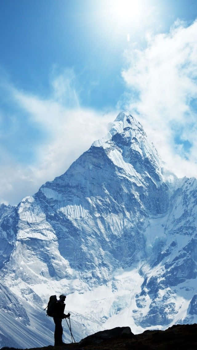 Winter Hiking 桌布 壁紙 — 一個孤獨的遠足者拿著登山杖，在明亮的藍天和縷縷白雲下，以剪影形式矗立在一座高聳的雪山前。