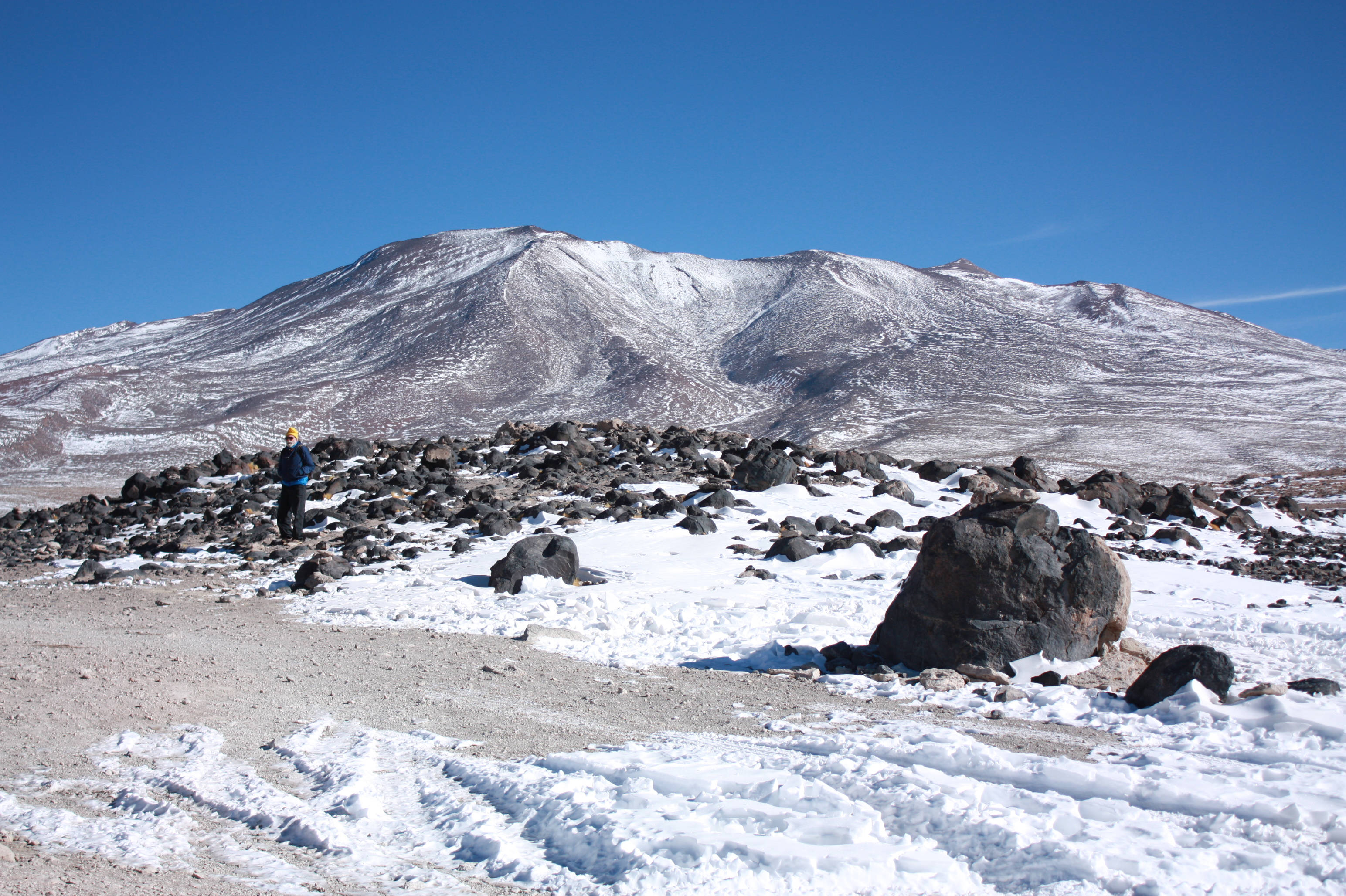 Bolivia 桌布 壁紙 — 一個孤獨的遠足者穿著藍色外套和黃色帽子，站在白雪覆蓋的岩石斜坡上，頭頂是廣闊清澈的藍天，面對著積雪的山脈。