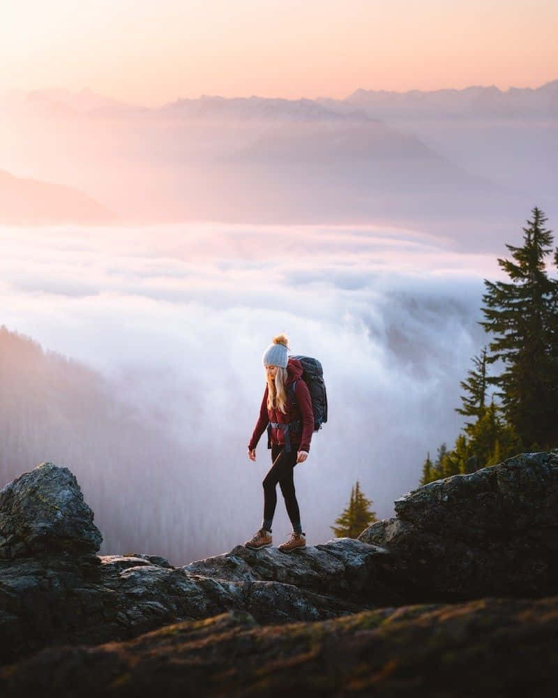 Winter Hiking 桌布 壁紙 — 一個孤獨的遠足者背著背包站在岩石懸崖頂上，俯瞰日出時的霧海，遠山沐浴在柔和的粉紅色光中。