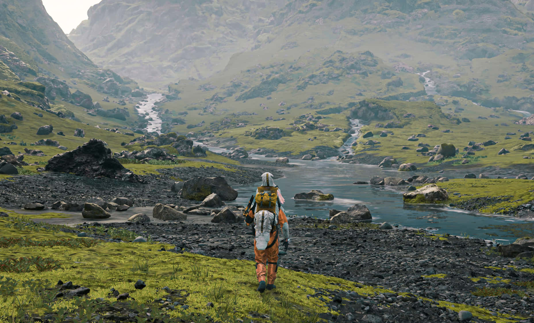 Landscape Death Stranding Wide 壁紙 — 一個孤獨的遠足者背著色彩繽紛的背包，沿著岩石河岸穿過廣闊的綠色山谷，兩旁是霧氣繚繞的山脈。