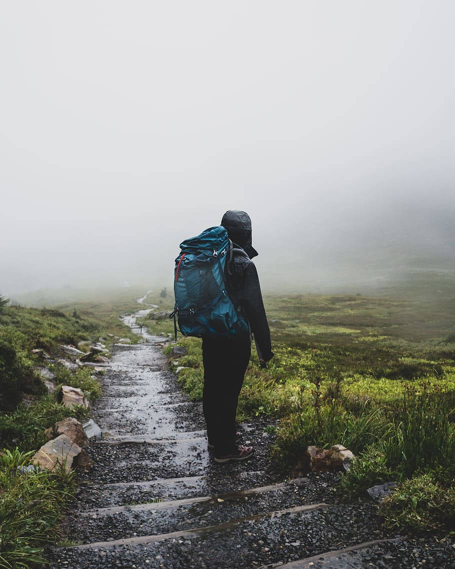 Hiking 桌布 壁紙 — 一個孤獨的遠足者背著藍色背包，站在一條濕滑的石徑上，石徑消失在濃霧籠罩的山巒中。
