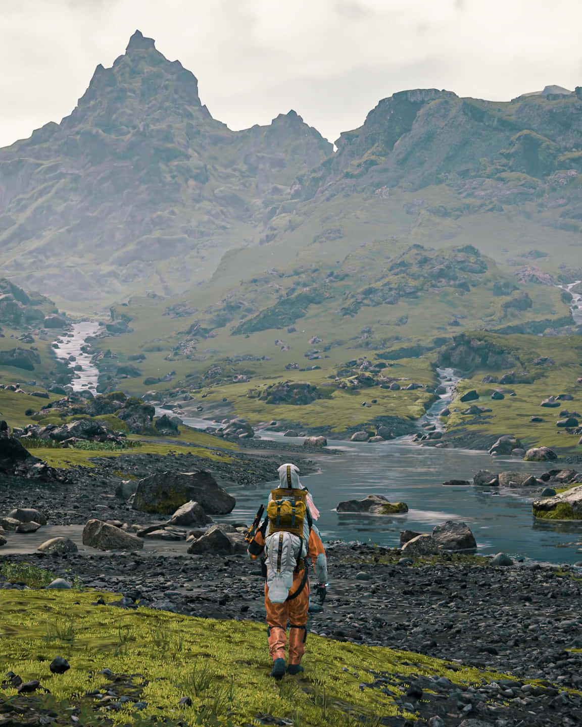 Land Scape Death Stranding 壁紙 — 一個孤獨的遠足者背著黃色背包，沿著長滿苔蘚的河岸，走向陰天天空下高聳的綠色山脈。