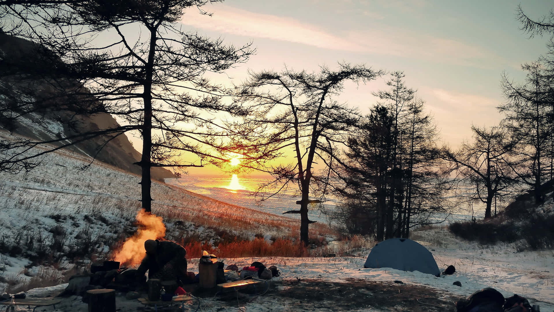 Camp 桌布 壁紙 — 一個孤獨的露營者在帳篷旁照料營火，夕陽落在由光禿樹木框住的雪山谷上。