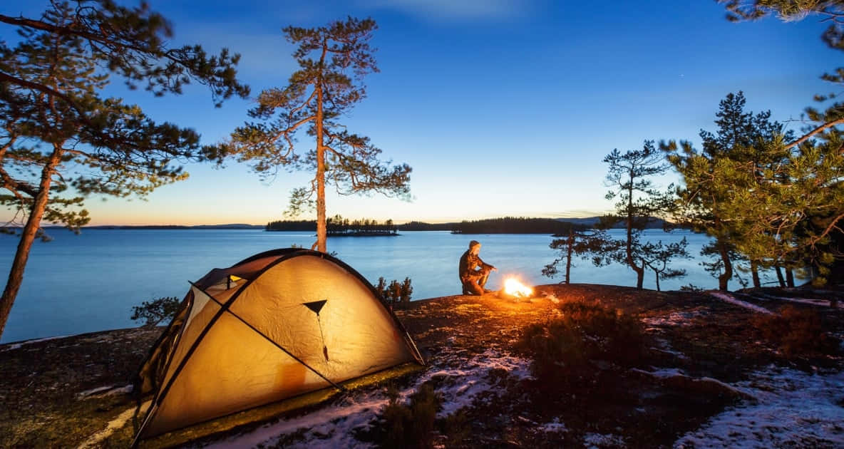 Fall Camping 桌布 壁紙 — 一個孤獨的露營者坐在帳篷旁邊發光的營火旁，在雪覆蓋的湖岸上，暮色中松樹在深藍色天空的襯托下形成剪影。