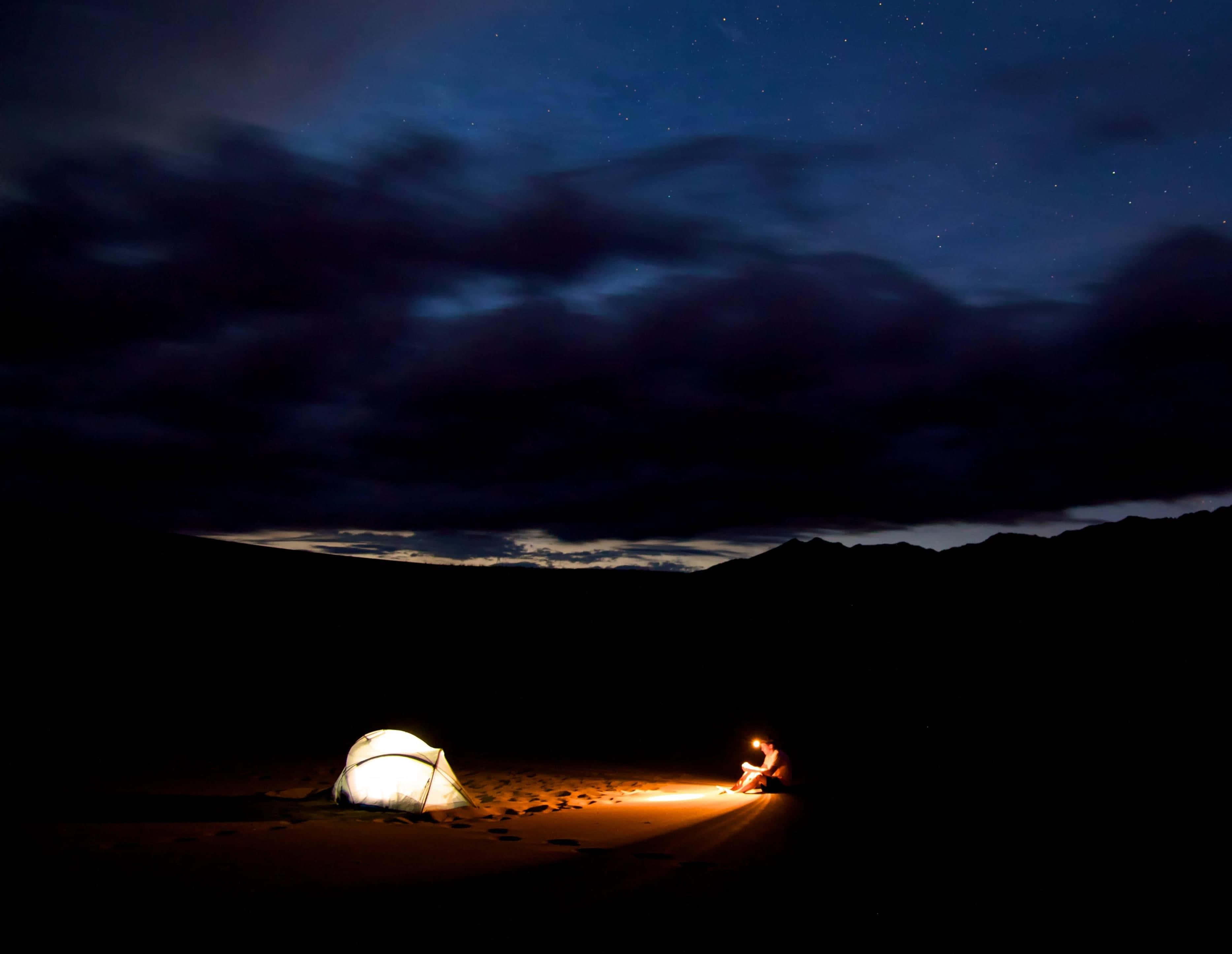 Black Clouds 桌布 壁紙 — 一個孤獨的露營者坐在發光的營火旁，在繁星點點的夜空下，附近有一個亮著的帳篷。