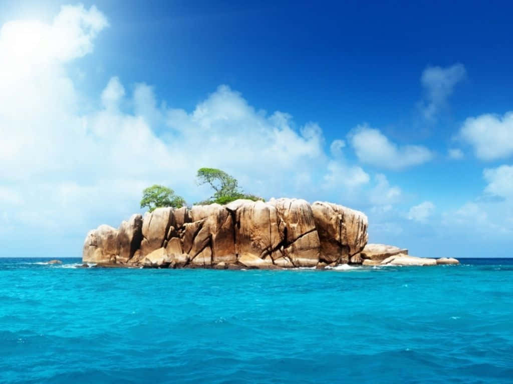島嶼 桌布 壁紙 — 一個孤立的岩石島嶼上長滿了綠樹，從充滿活力的碧綠海水中升起，上方是明亮的藍天和蓬鬆的白雲。