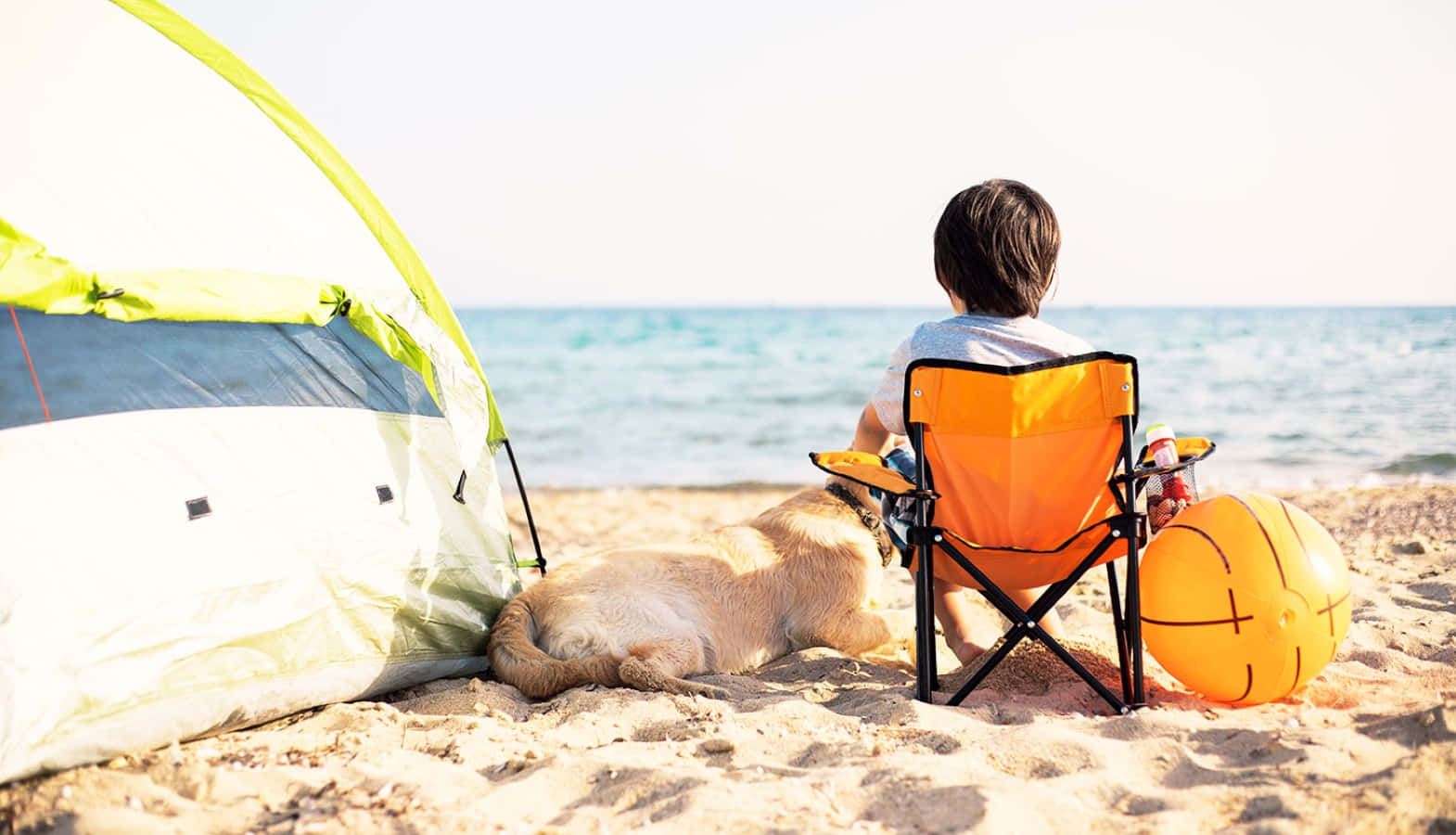 Beach Camping 桌布 壁紙 — 一個孩子和一隻狗在沙灘上，在帳篷和橙色椅子旁邊放鬆，在明亮的陽光下凝視著海洋。