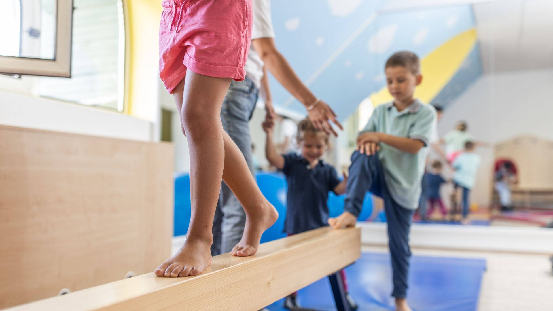 Playground 桌布 壁紙 — 一個孩子在明亮的室內遊戲室赤腳平衡在木樑上，而成人和其他孩子在附近進行體能活動。
