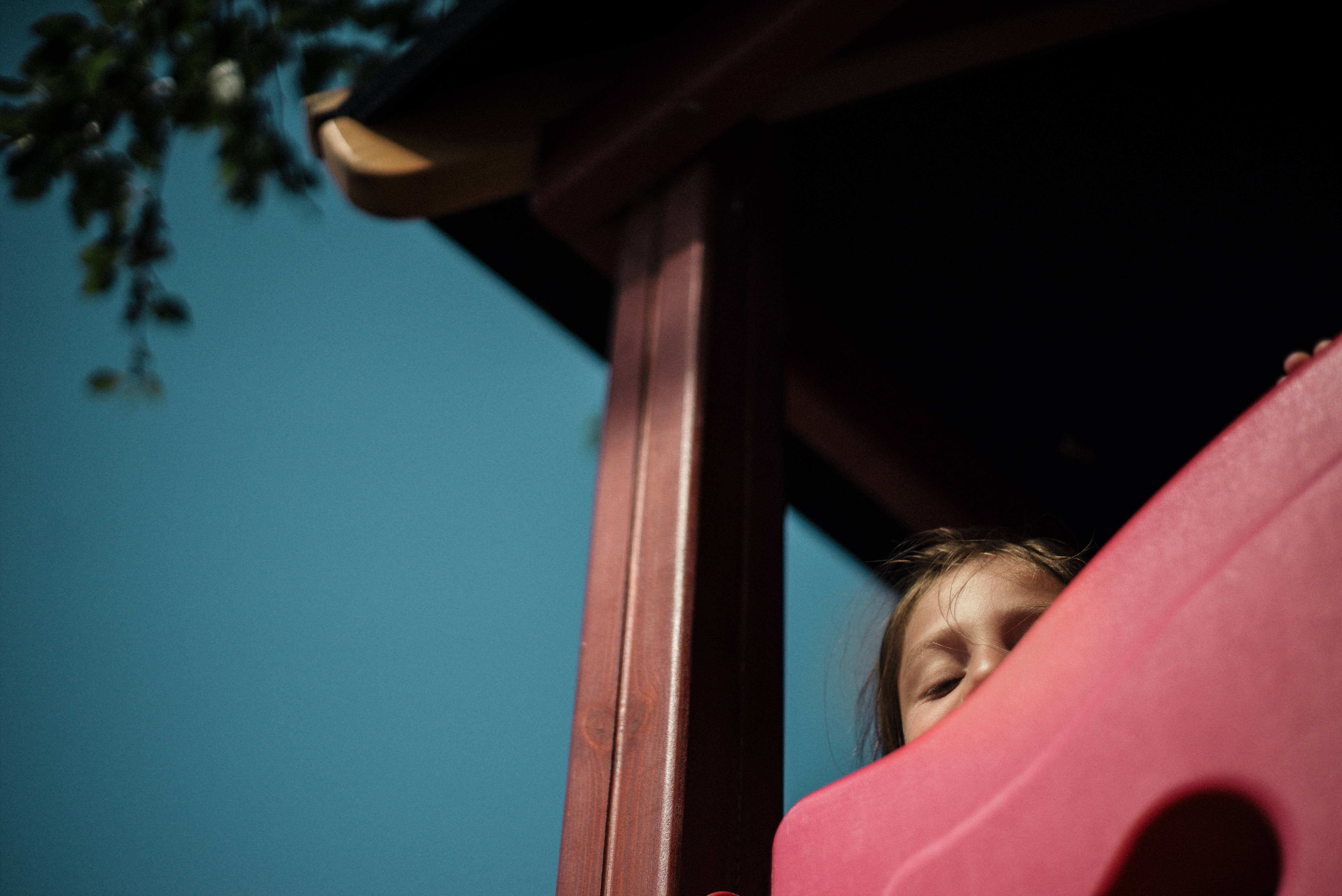 Playground 桌布 壁紙 — 一個孩子在晴朗的藍天下，好奇地從紅色塑膠溜滑梯後面探出頭來。
