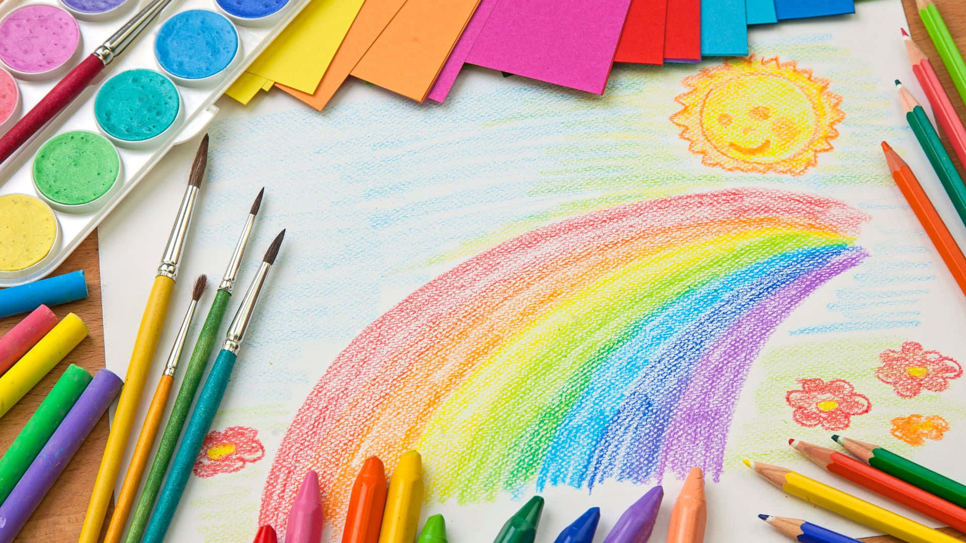 小孩動圖 桌布 壁紙 — 一個孩子在木桌上用彩色蠟筆畫的笑臉太陽和彩虹，周圍環繞著美術用品。