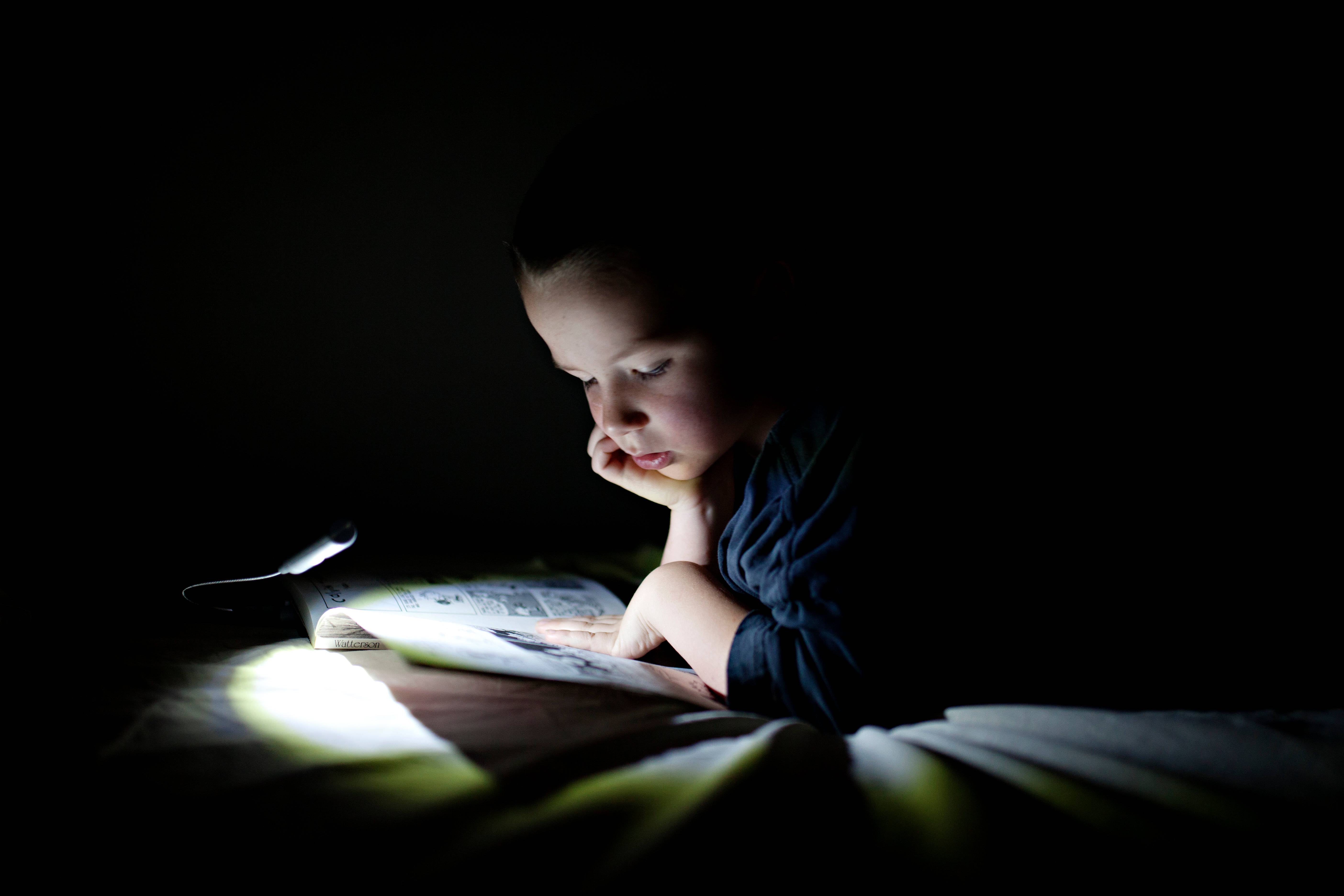 Study 桌布 壁紙 — 一個孩子在黑暗的房間裡，在明亮的手電筒下專注地閱讀一本書。