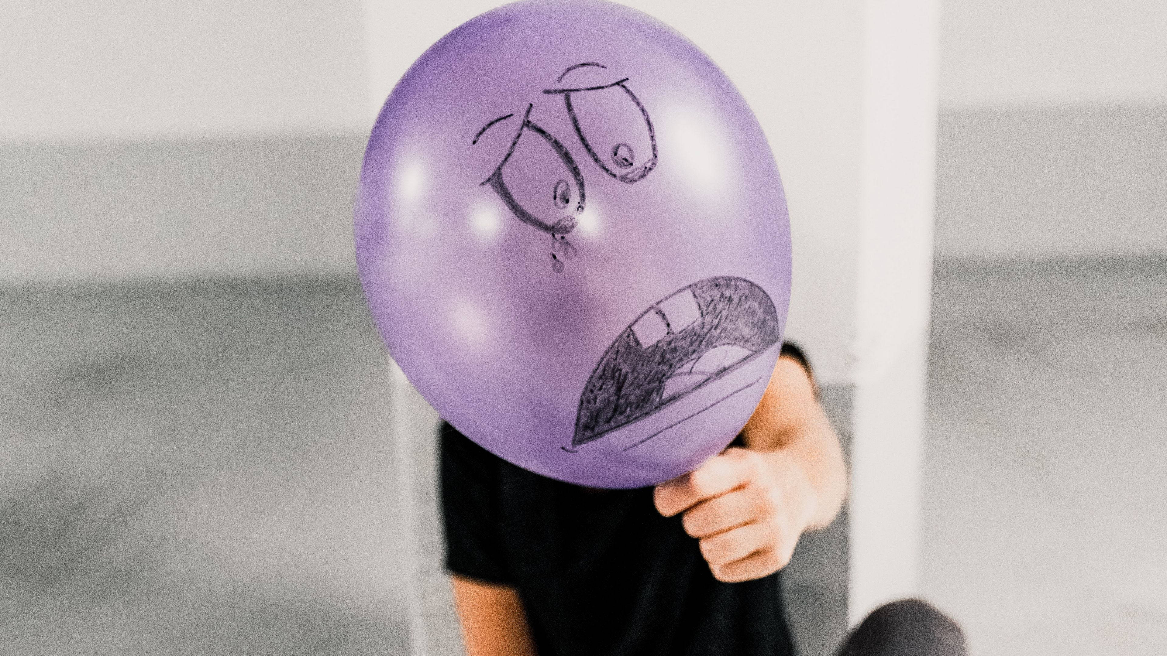 Balloon 桌布 壁紙 — 一個孩子拿著一個紫色氣球，上面畫著悲傷的臉，遮住了他們的頭，在一個簡約的室內空間。