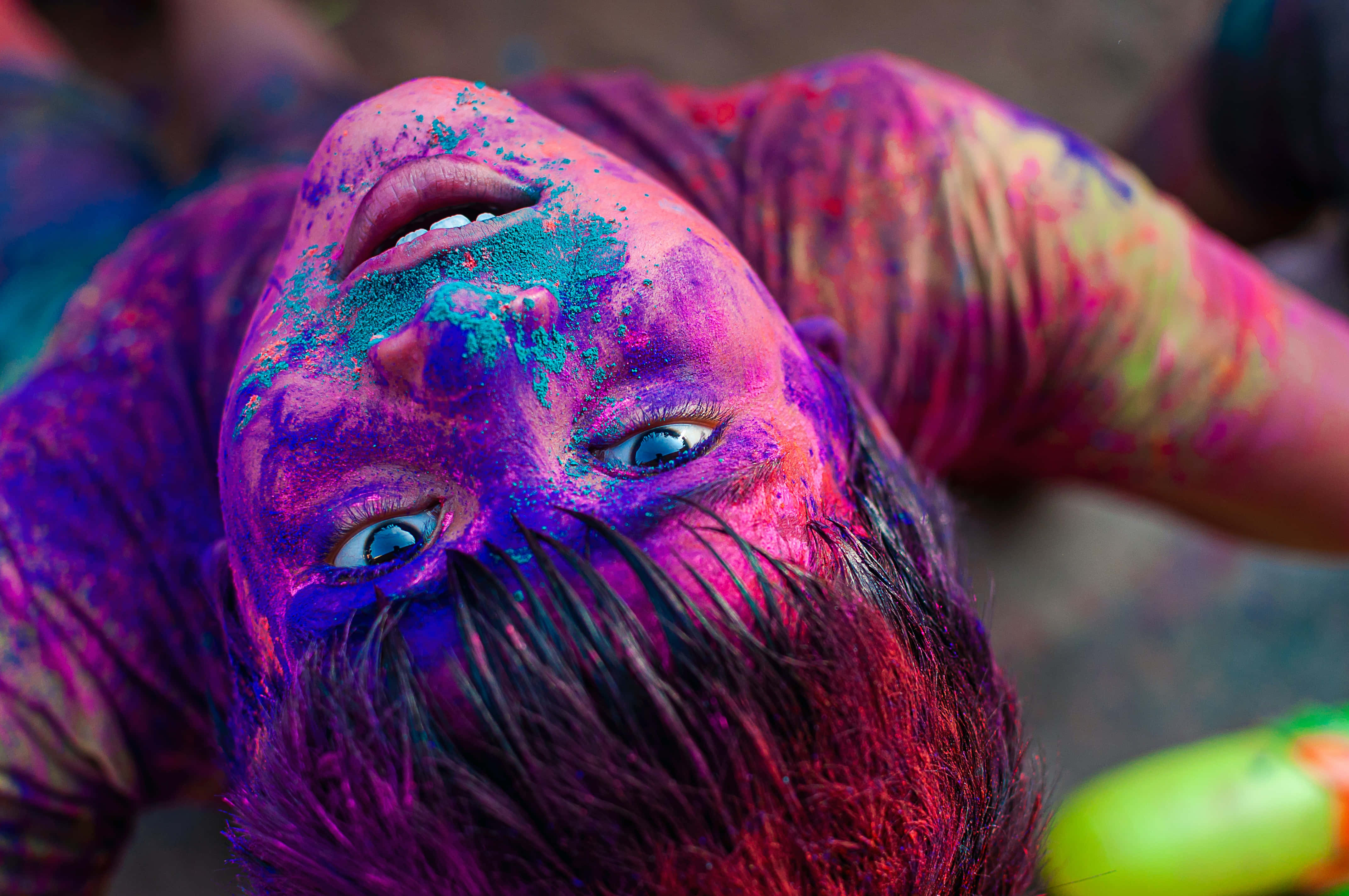 Holi 桌布 壁紙 — 一個孩子的臉和上半身被鮮豔的紫色、粉紅色和藍綠色粉末覆蓋，抬起頭來，有著明亮的藍色眼睛。