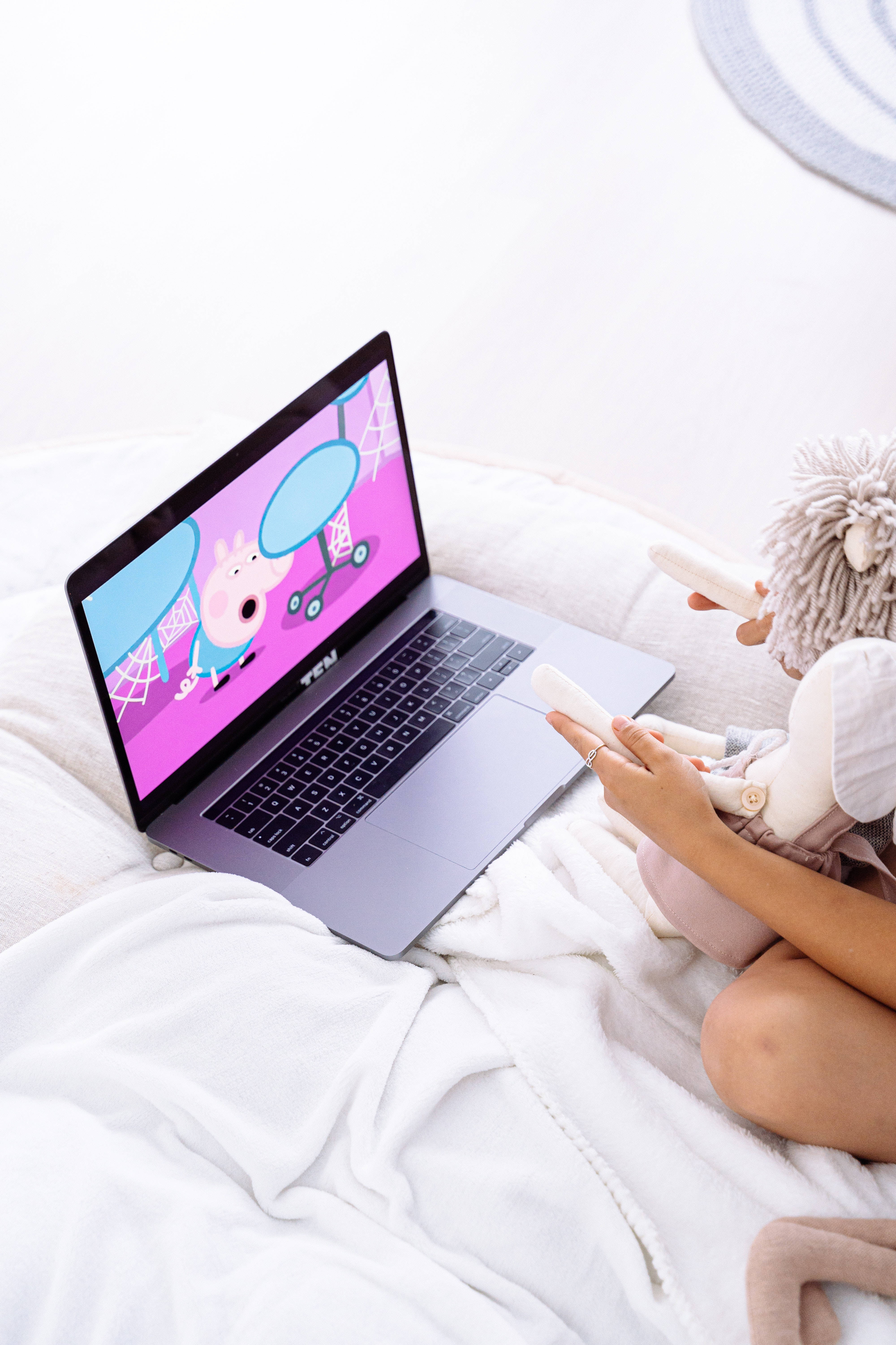 aesthetic tumblr laptop 桌布 壁紙 — 一個孩子舒適地坐在床上，用筆記型電腦播放Peppa Pig，同時抱著毛絨玩具和遙控器。
